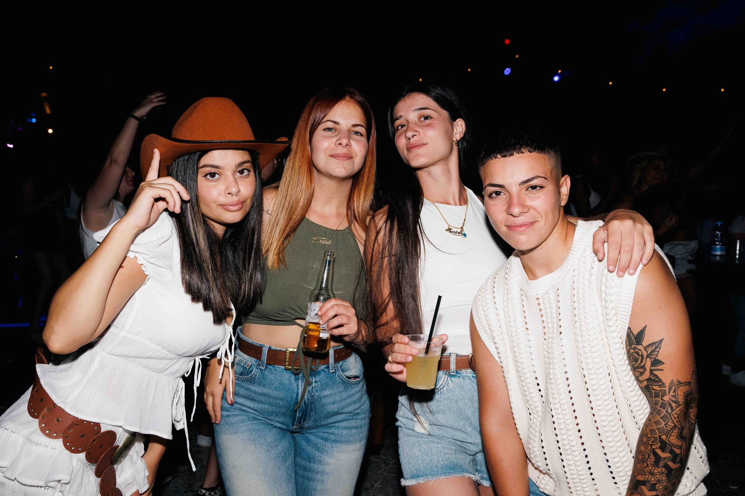 Four young women at a party, smiling and holding drinks, with dark background and faint colorful lights.