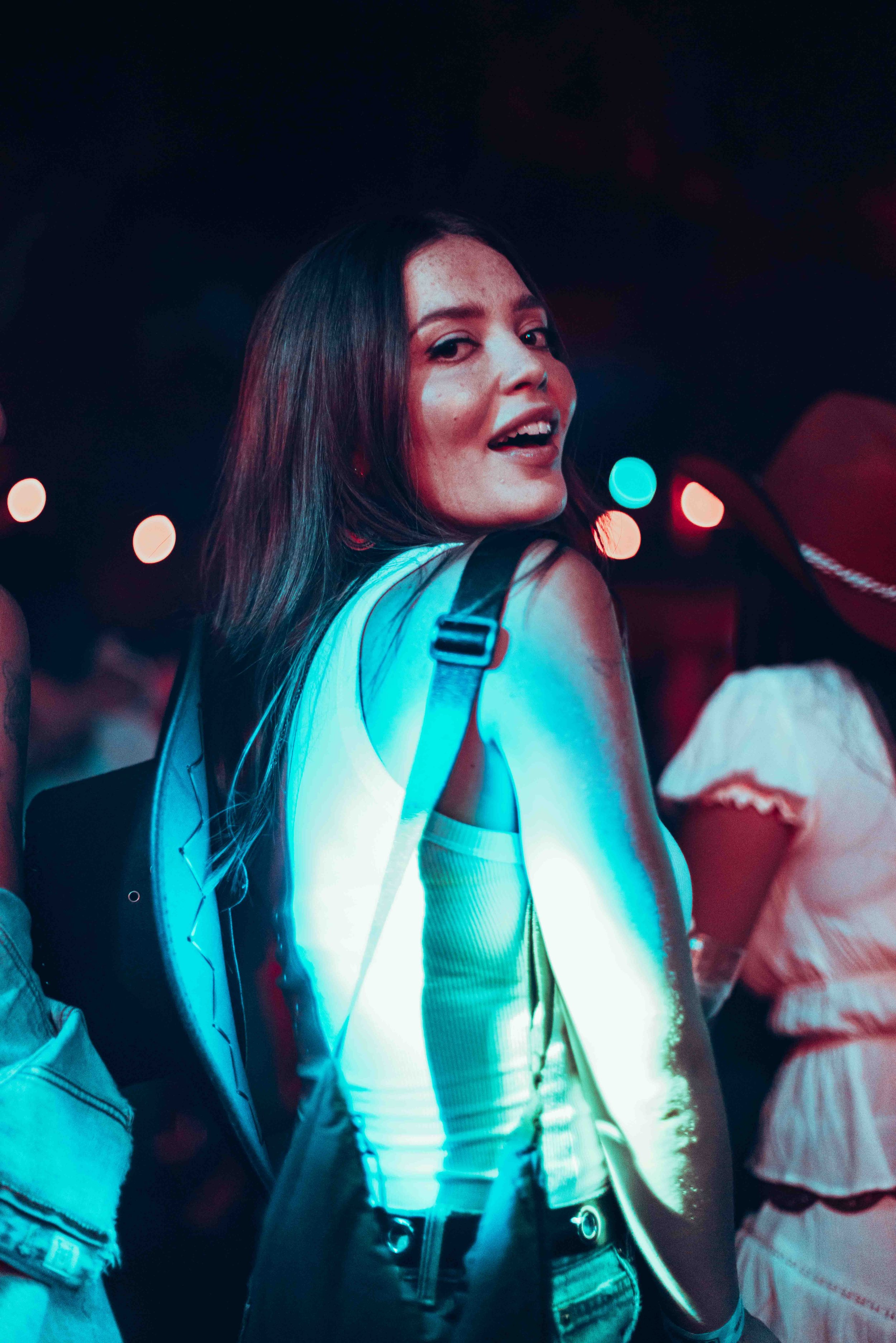A young woman with long dark hair smiling and looking over her shoulder at night, illuminated by colorful city lights, standing beside other people in a social setting.