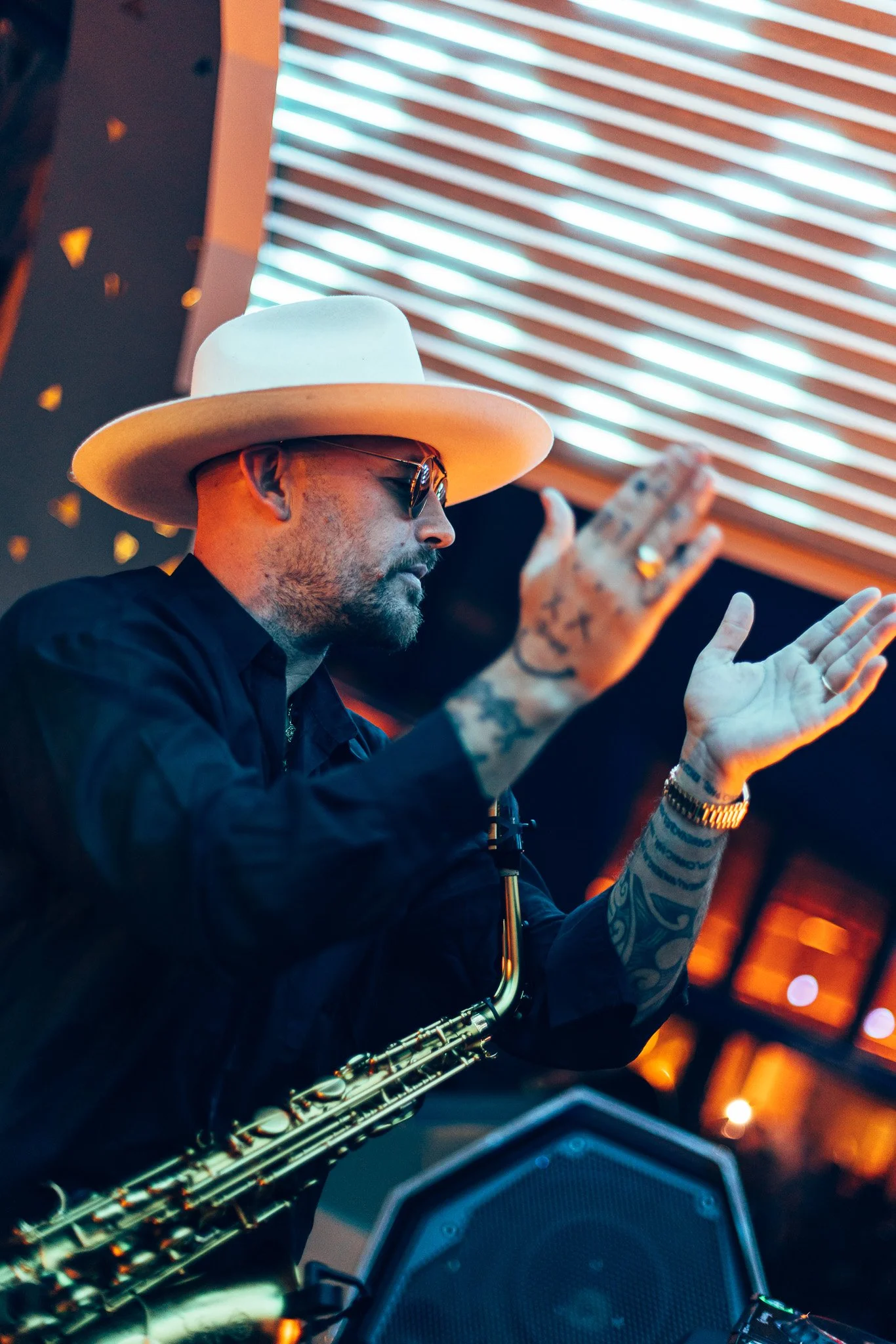 A man with tattoos wearing glasses and a white hat playing a saxophone indoors, with colorful lighting and a modern background.