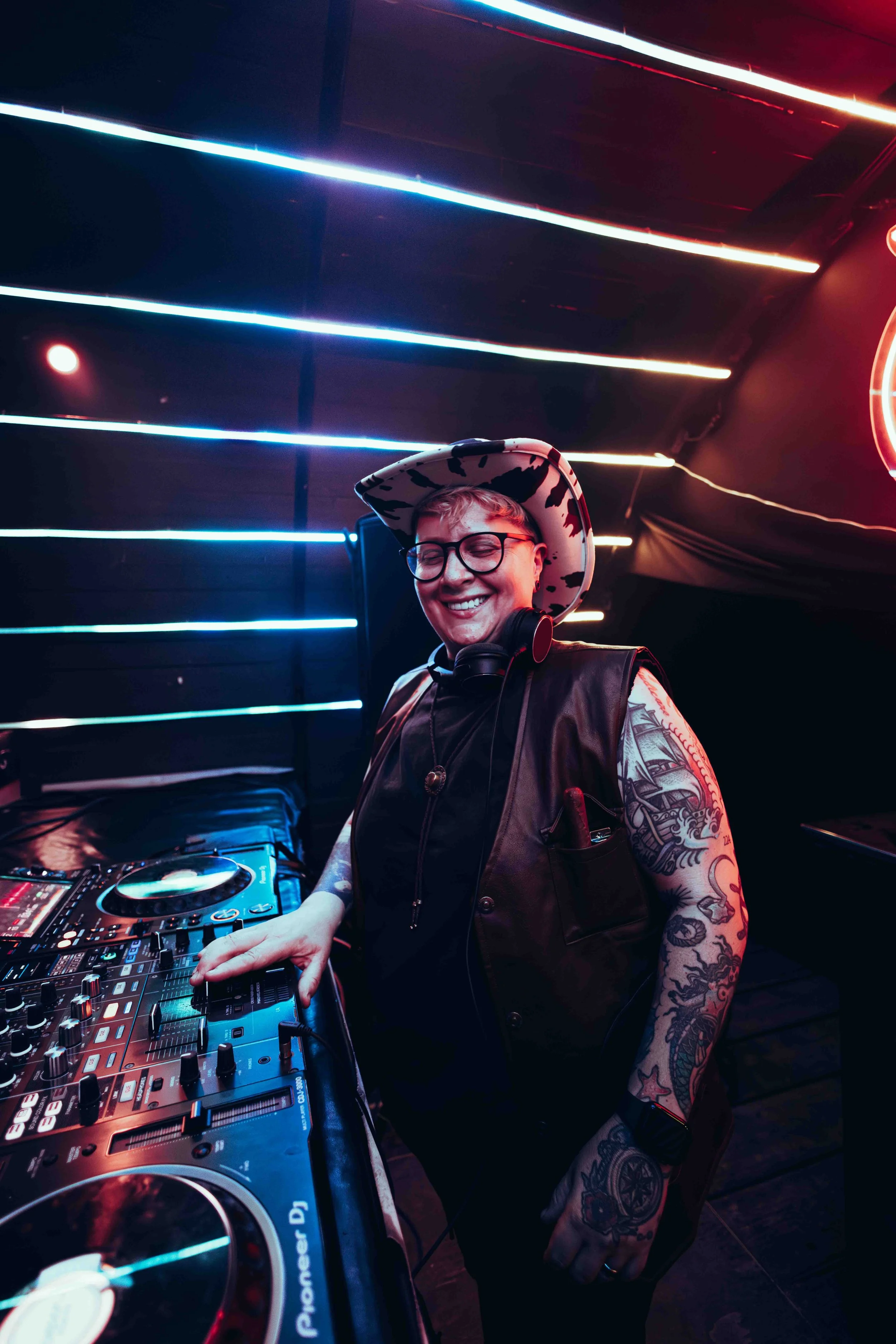 Person smiling with tattoos, wearing glasses and a hat, DJing at a nightclub with colorful LED lights.