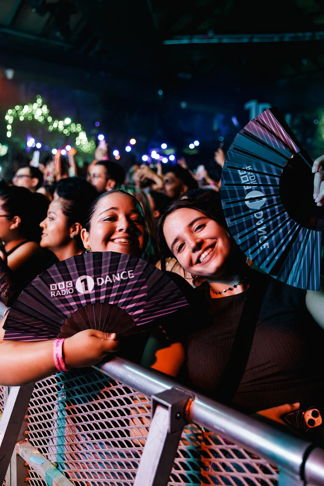 Group of smiling young women at a concert waving fans with BBC Radio 1 Dance logo, colorful lighting, crowd in background.