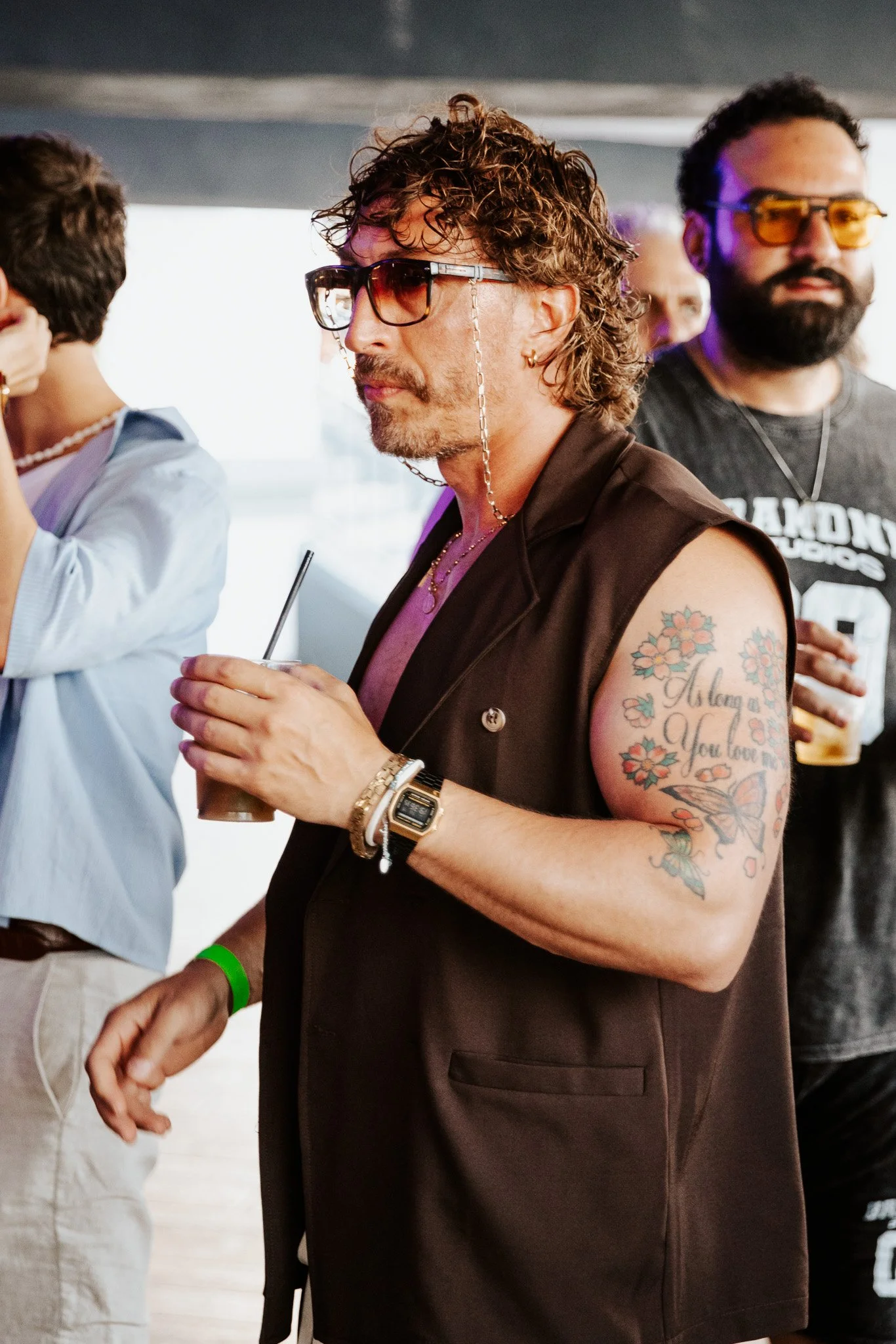 Man with curly hair and tattoos on his arm, wearing sunglasses and a sleeveless vest, holding a drink with a straw at what appears to be a social gathering or event.