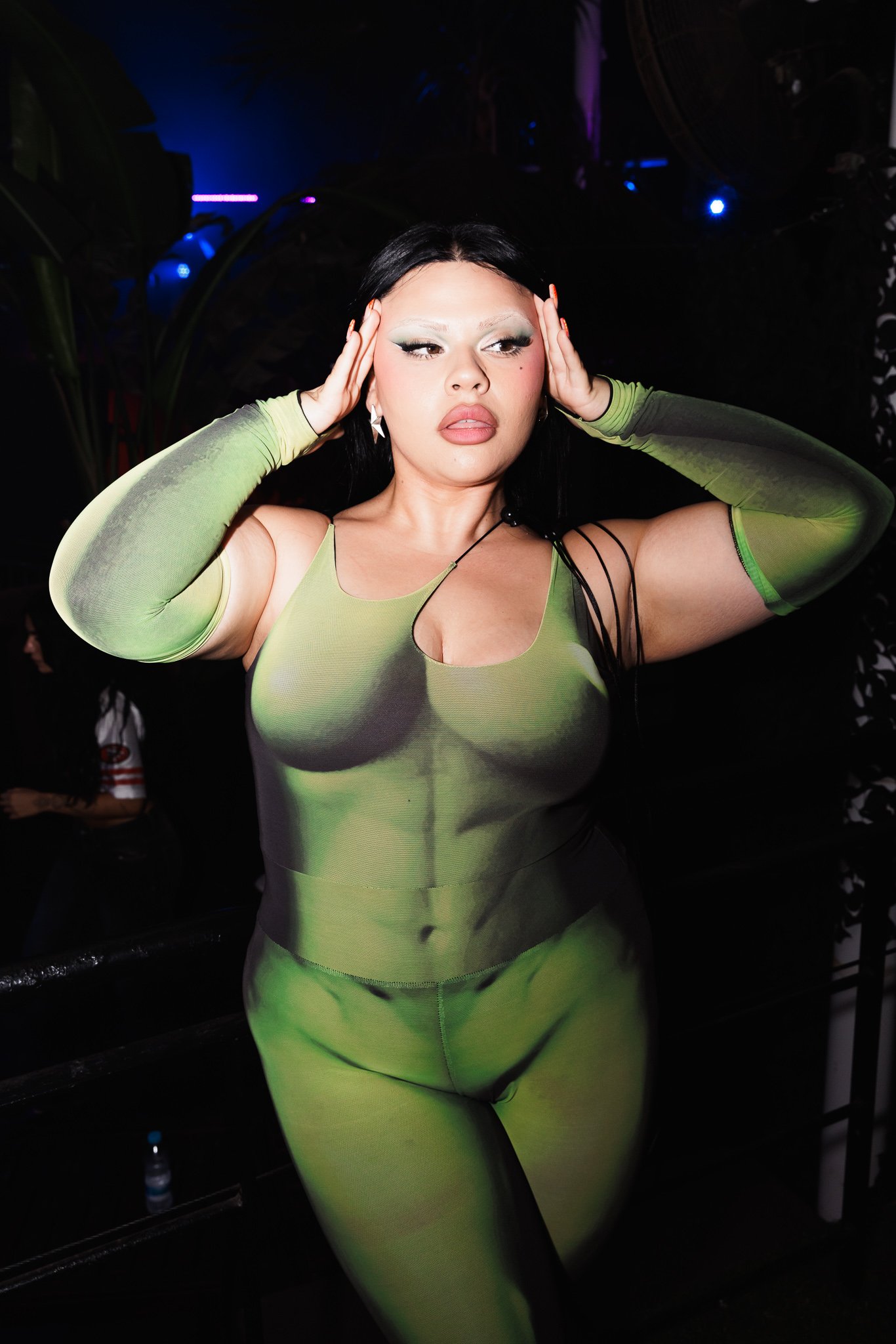 A woman with long black hair is holding her hands to her head, wearing a neon green, sheer dress with matching sleeves, in a dark setting with colorful lights.