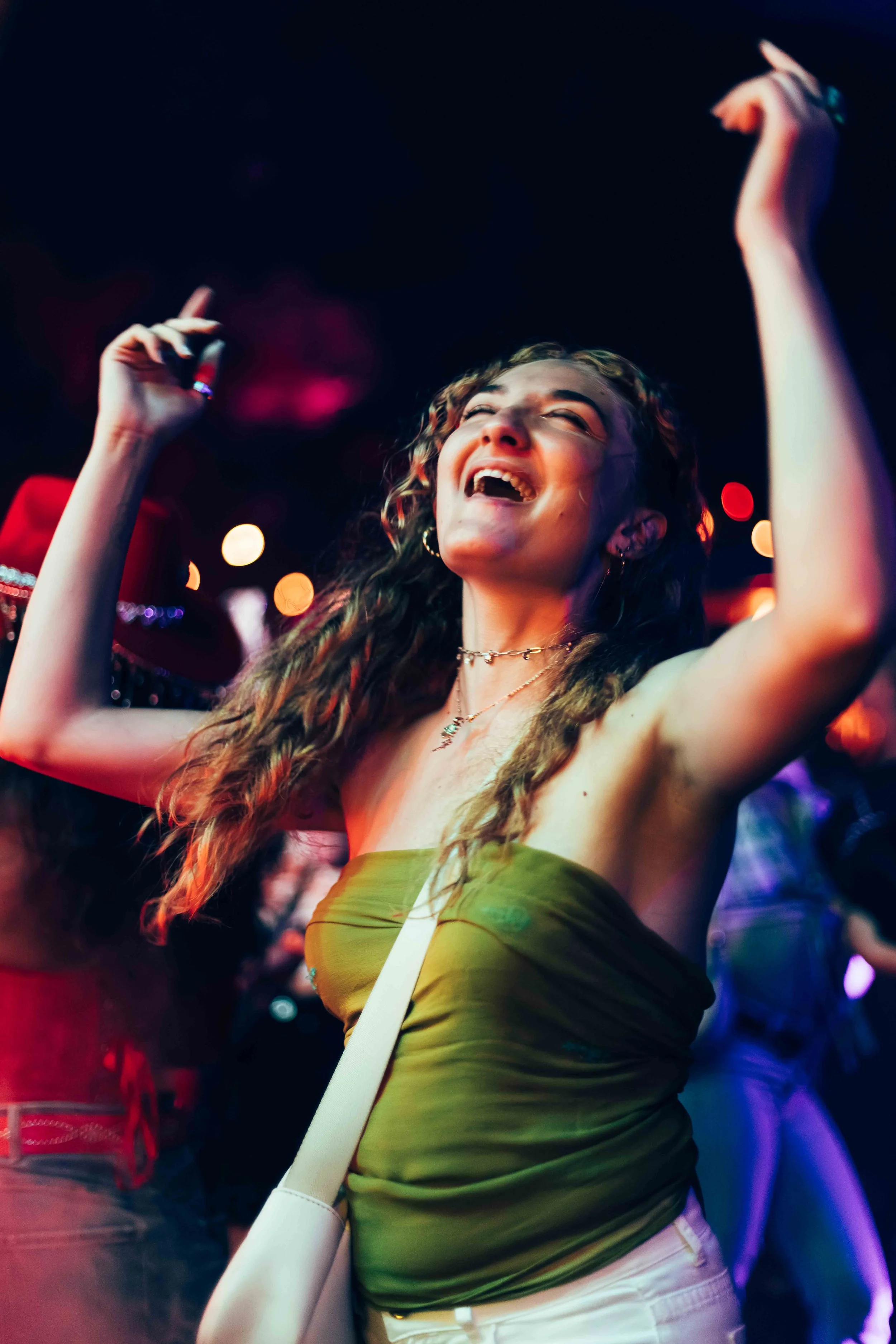 A joyful young woman dancing in a club with colorful lights, wearing a green sleeveless top, white pants, and a white purse.