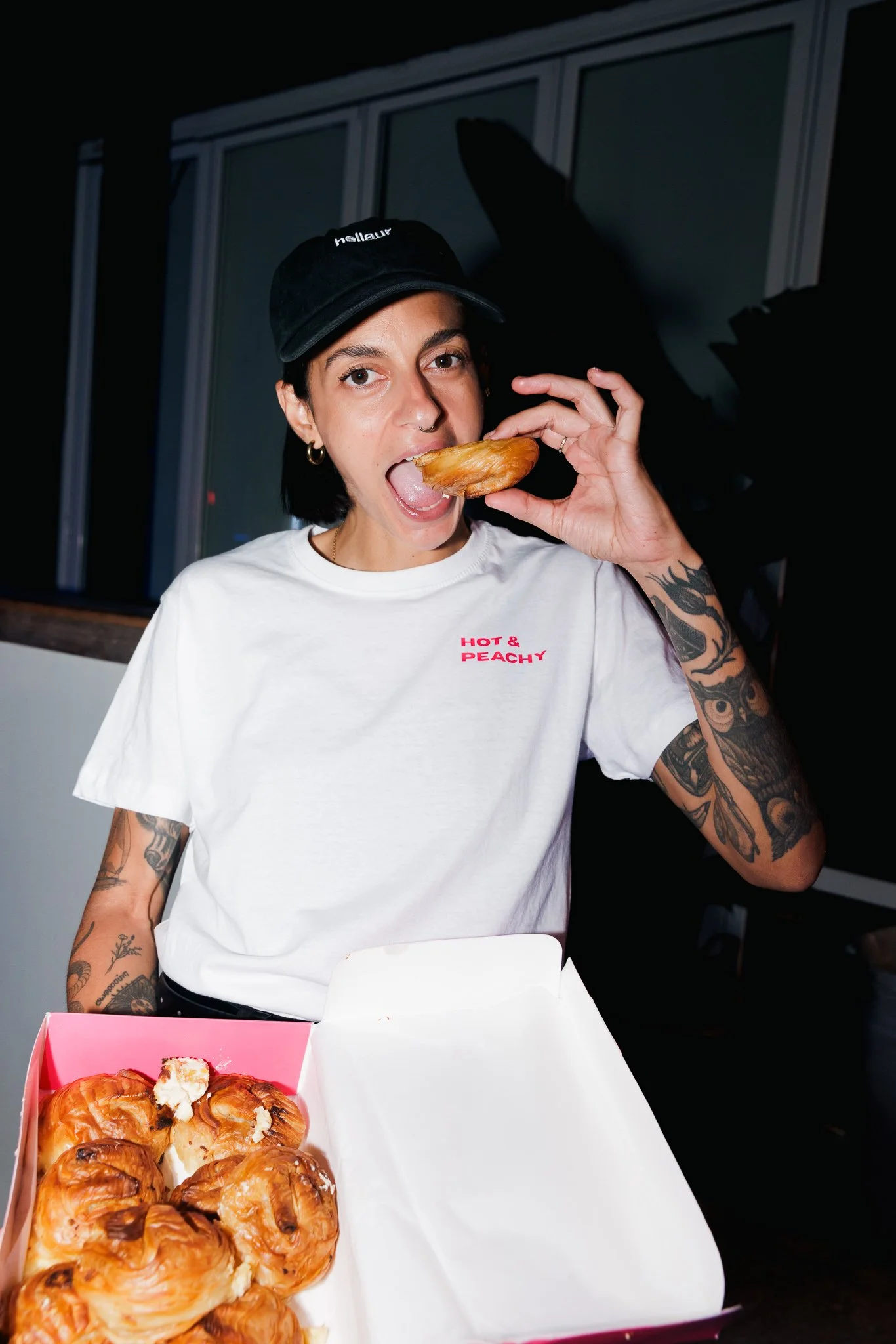 A woman with tattoos on her arms and ear piercings is wearing a black baseball cap and a white t-shirt that says 'Hot & Peachy'. She is holding a pastry and about to eat it, with a box of croissants and pastries in front of her.