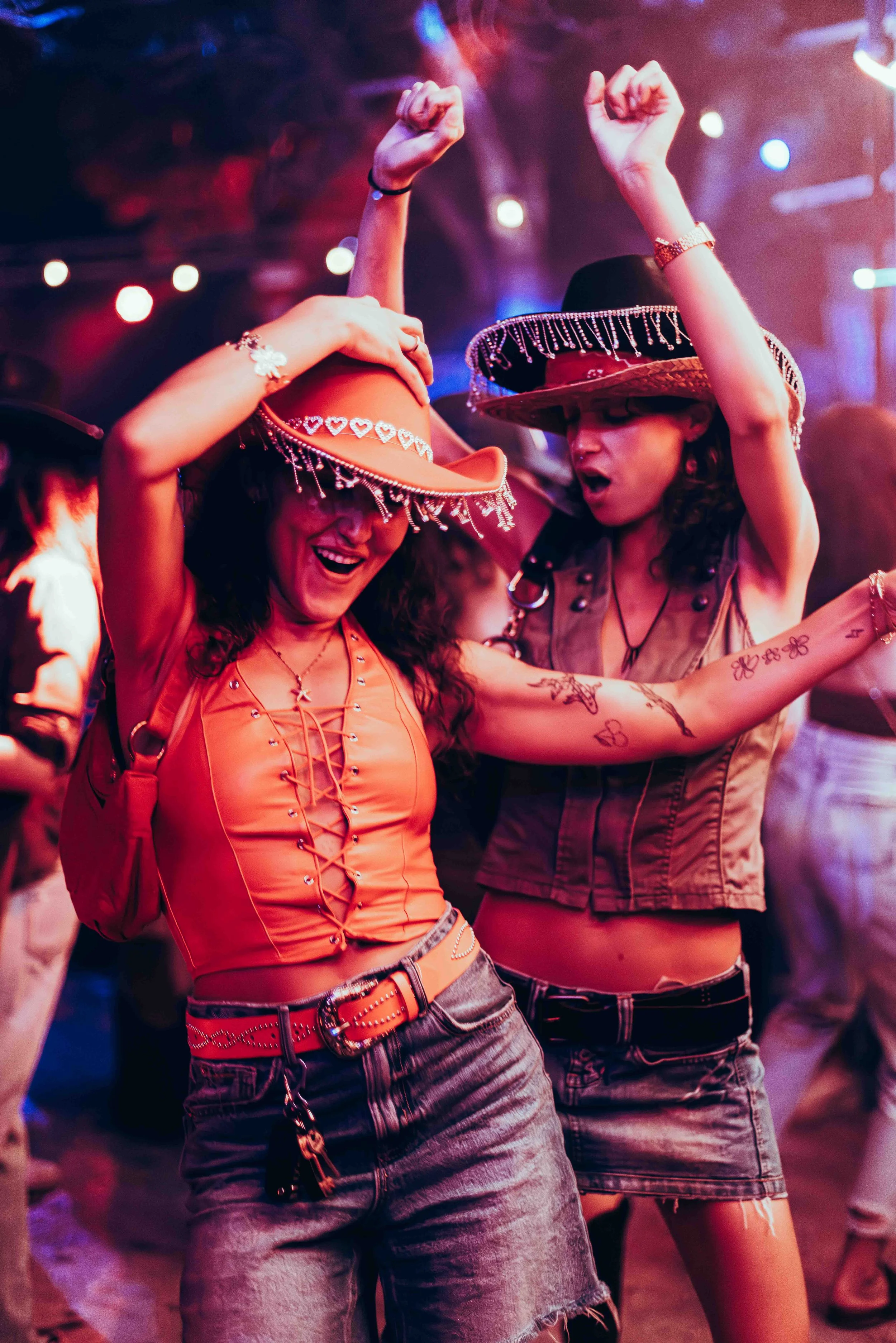 Two women at a party or dance event, both wearing cowboy hats with decorative beads. The woman on the left has long dark hair, glasses, and is smiling while dancing, wearing a sleeveless orange top with lace-up front, and a red purse. The woman on th