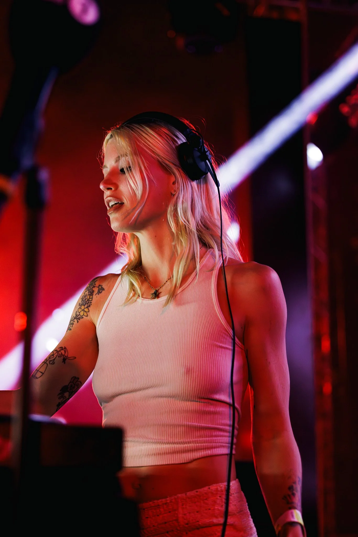A woman with blonde hair wearing a light pink sleeveless top, headphones, and tattoos on her arm, appears to be DJing at a club or event with colorful lighting.