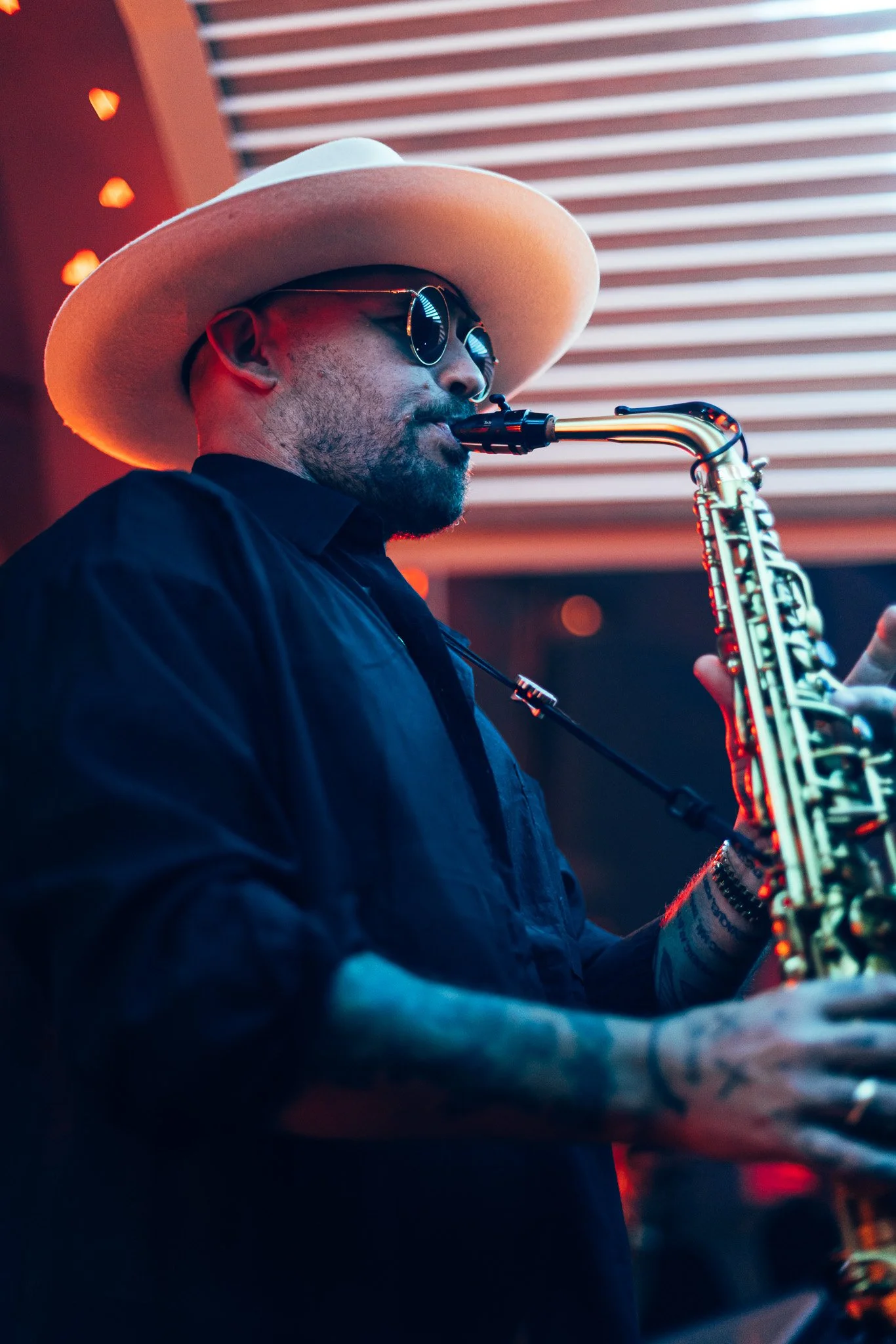 A man dressed in black, wearing sunglasses and a wide-brimmed hat, playing a saxophone in an indoor setting with red and warm lighting.