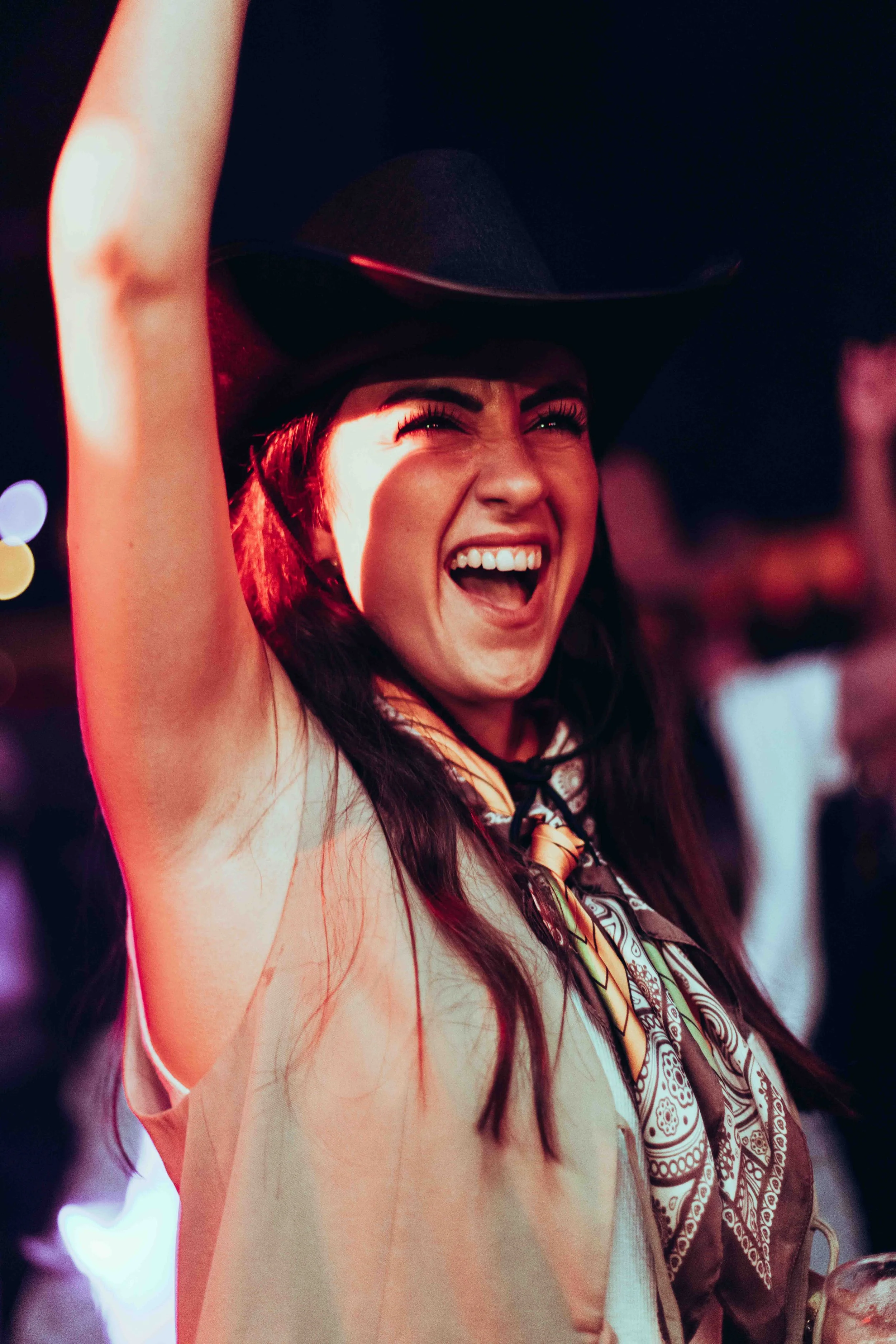A woman smiling with her mouth open, wearing a wide-brimmed black hat and a patterned bandana around her neck, raising her arm in a lively setting at night.