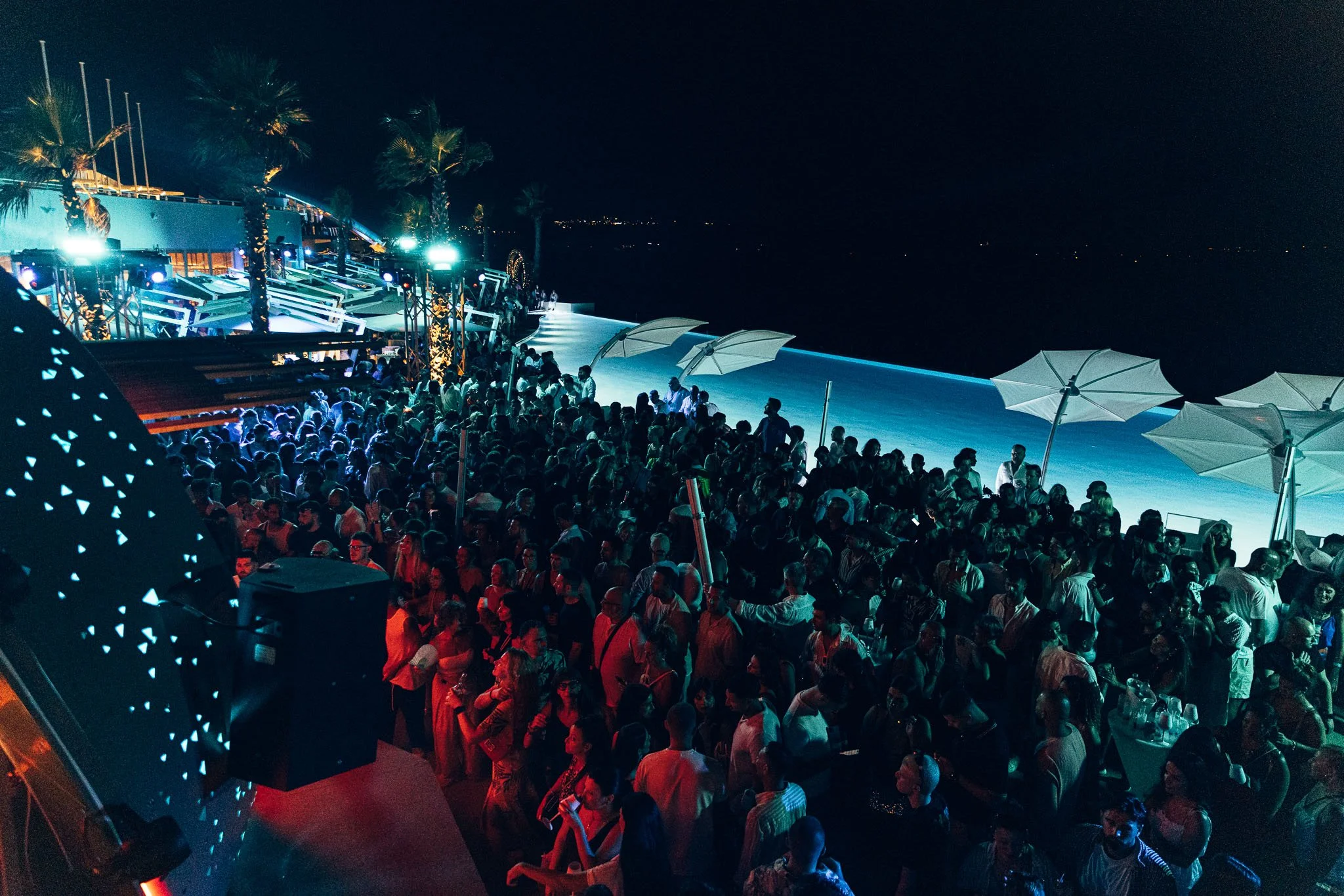 Nighttime scene at a crowded outdoor event or party with many people and large umbrellas, near a body of water, illuminated by colorful lighting.