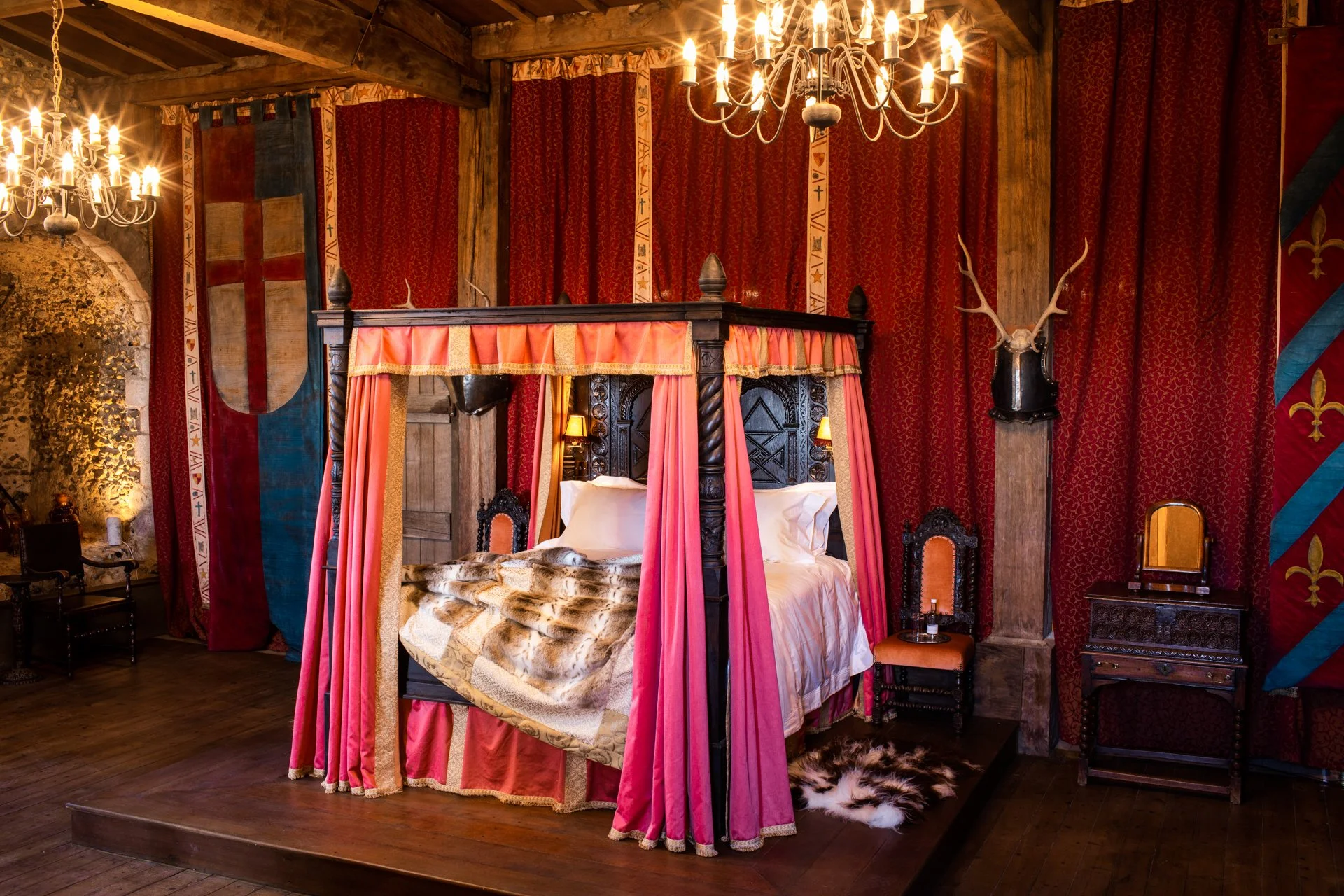 Medieval-themed bedroom with a canopy bed, red and gold drapes, antique furniture, chandeliers, and wall banners.