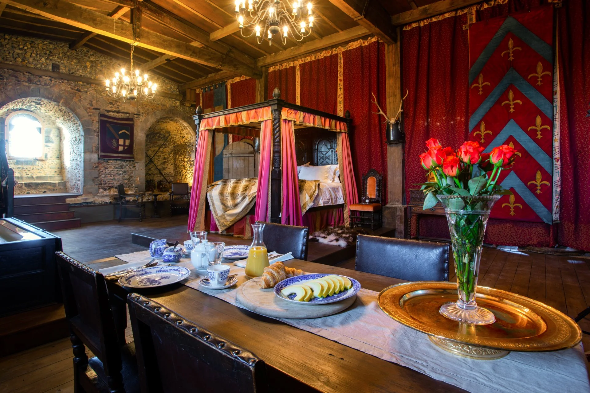 A medieval-style room with a four-poster bed draped in pink fabric, red curtains with heraldic patterns, stone walls, and wooden ceiling. A wooden dining table is set with a vase of red roses, breakfast items, and blue and white crockery. Chandeliers