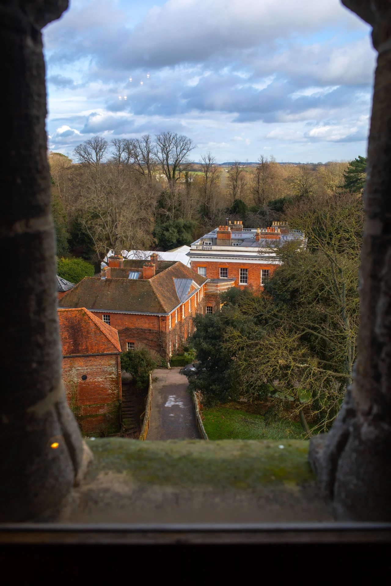 View from a stone window of a historic brick building surrounded by trees and a courtyard, under a cloudy sky.