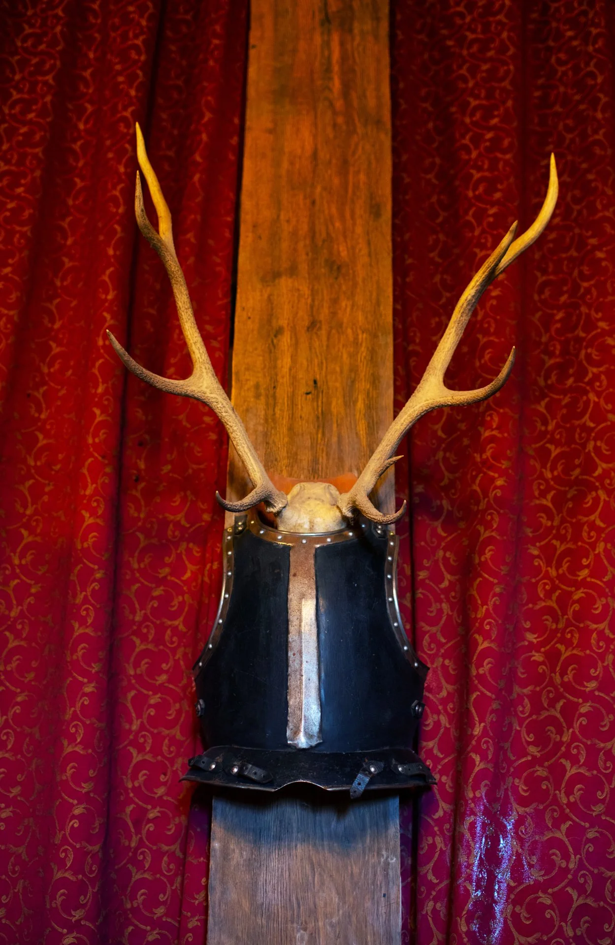 Antlers mounted on a medieval-style helmet, displayed on a wooden board against a red patterned curtain backdrop.