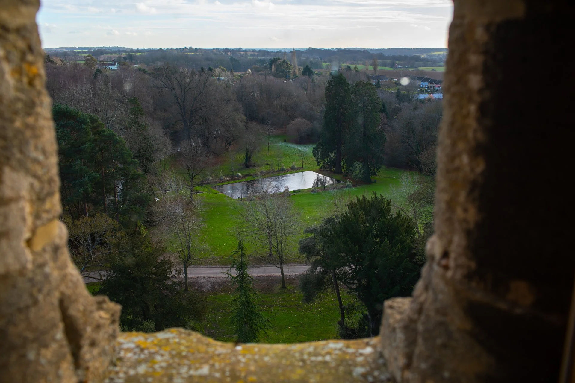 View through a castle window of a landscape with bare trees, grassy fields, and a rectangular pond, surrounded by forested hills in the distance.