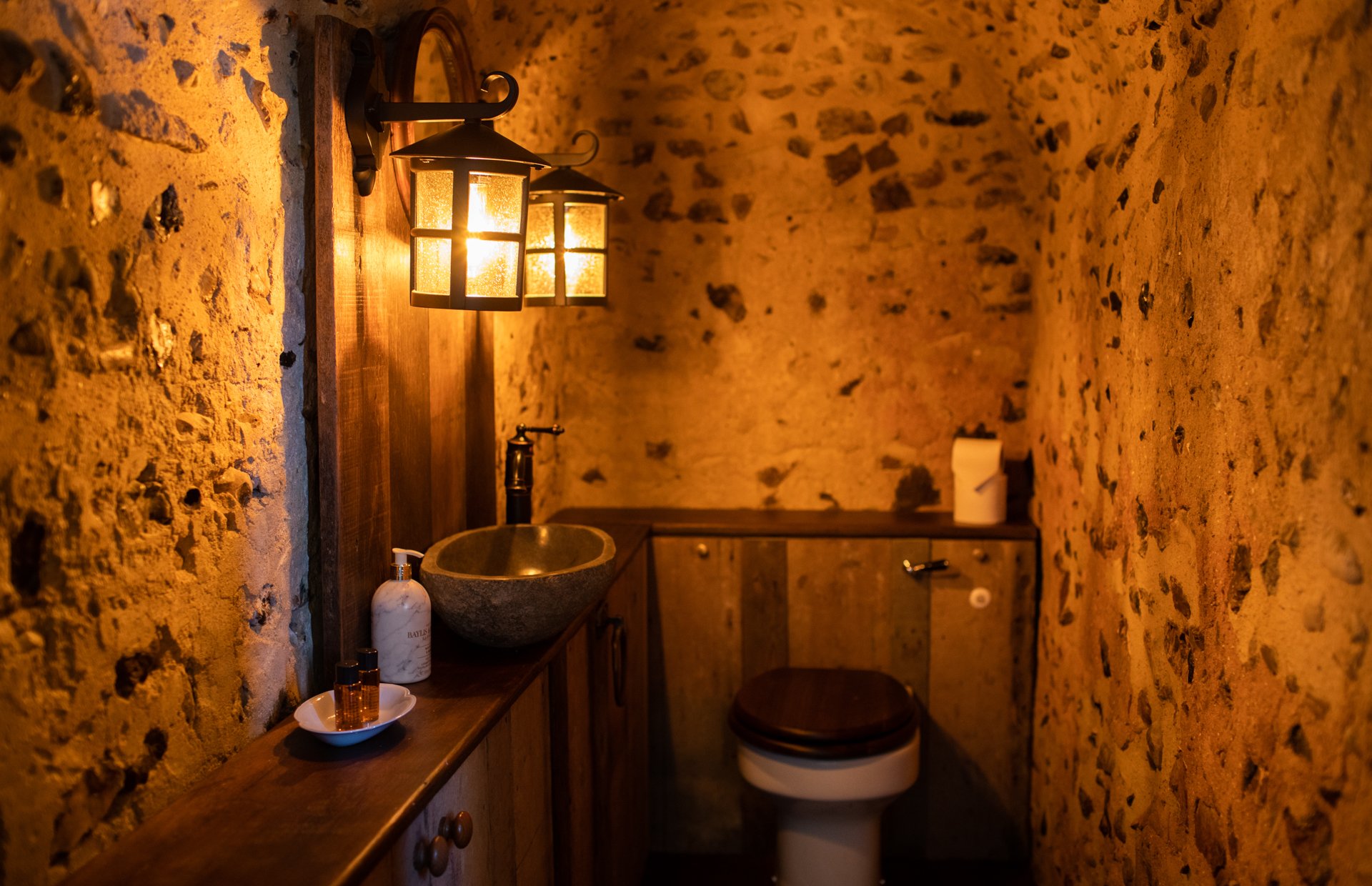 Rustic bathroom with stone walls, a round stone sink, wooden cabinets, a toilet, and warm lighting from lantern-style fixtures.