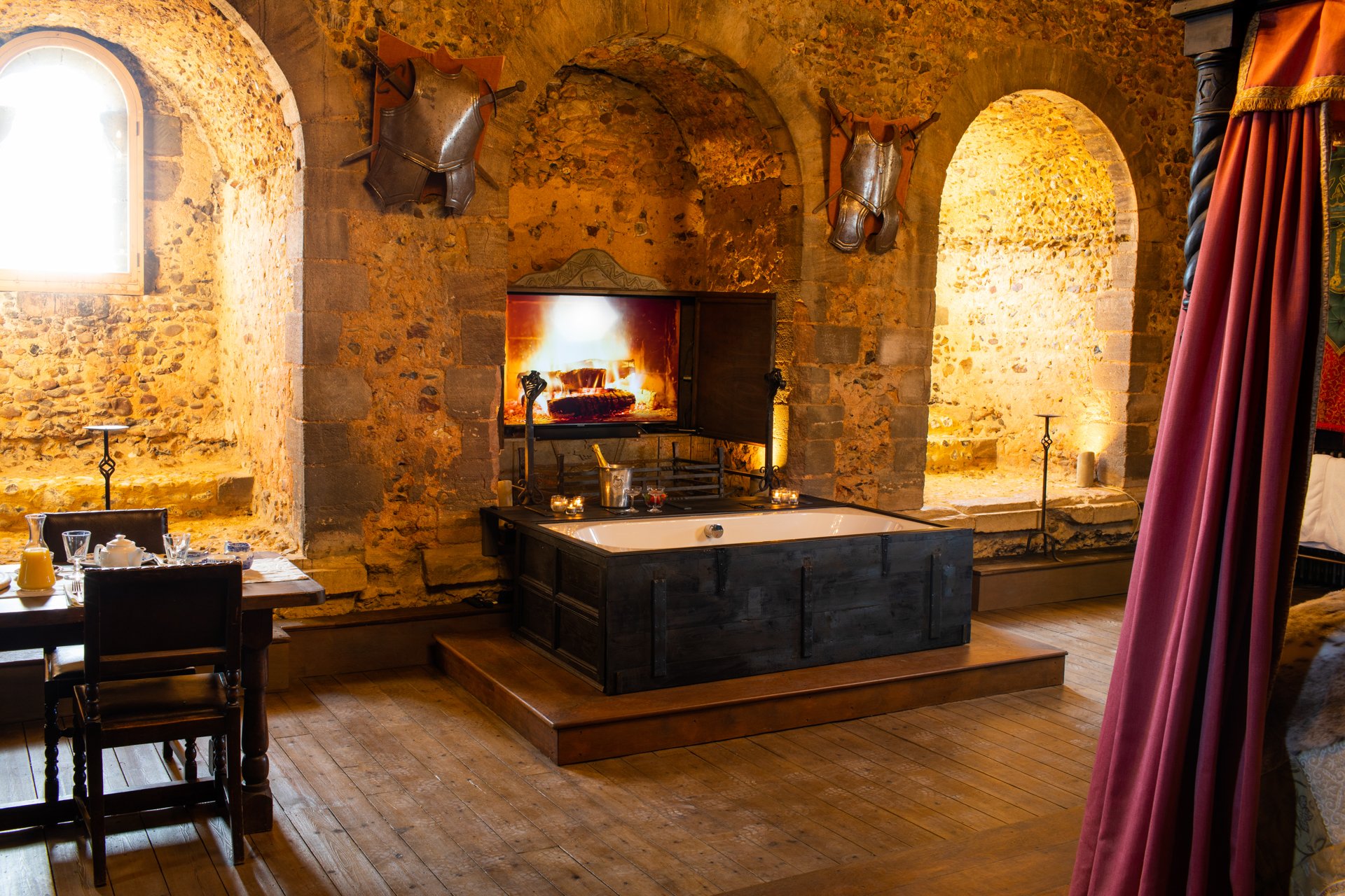 Medieval-style room with stone walls, a central bathtub, a fireplace screen with a fire image, a dining table, armored décor, and a draped four-poster bed.
