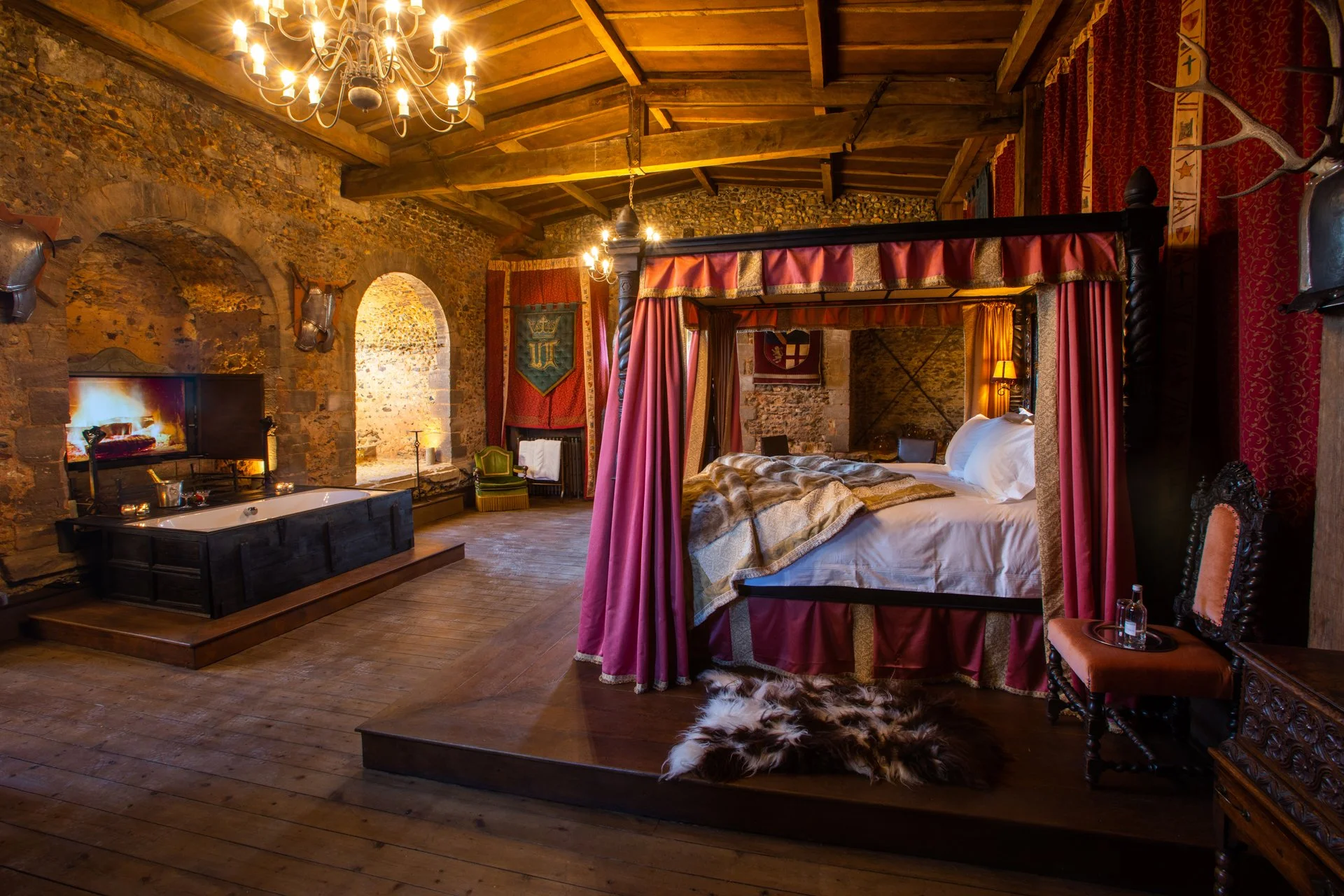 Medieval-style bedroom with a canopy bed, chandelier, stone walls, fireplace, bathtub, and various furnishings.