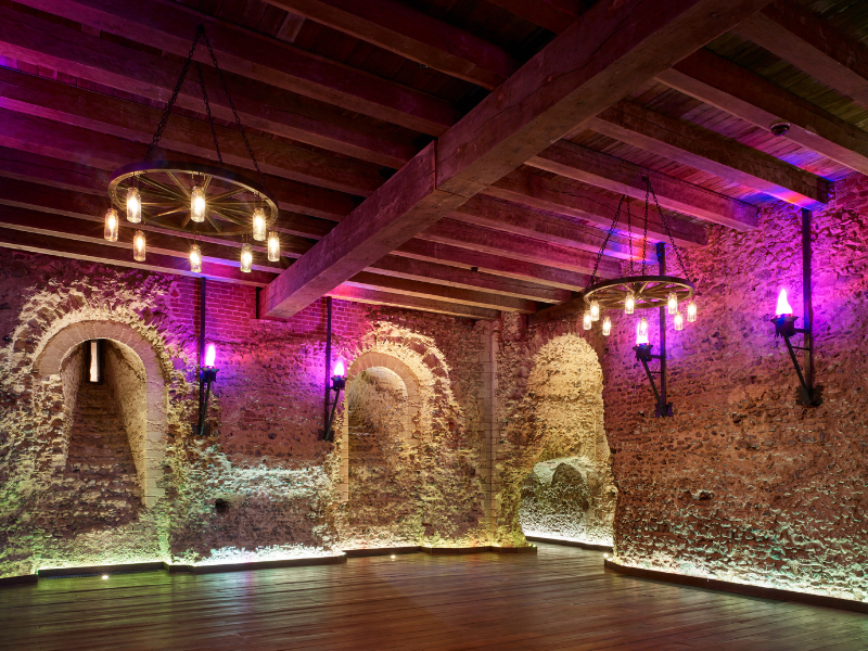 Rustic stone-walled interior with wooden beams, colorful wall lights, and hanging chandeliers.
