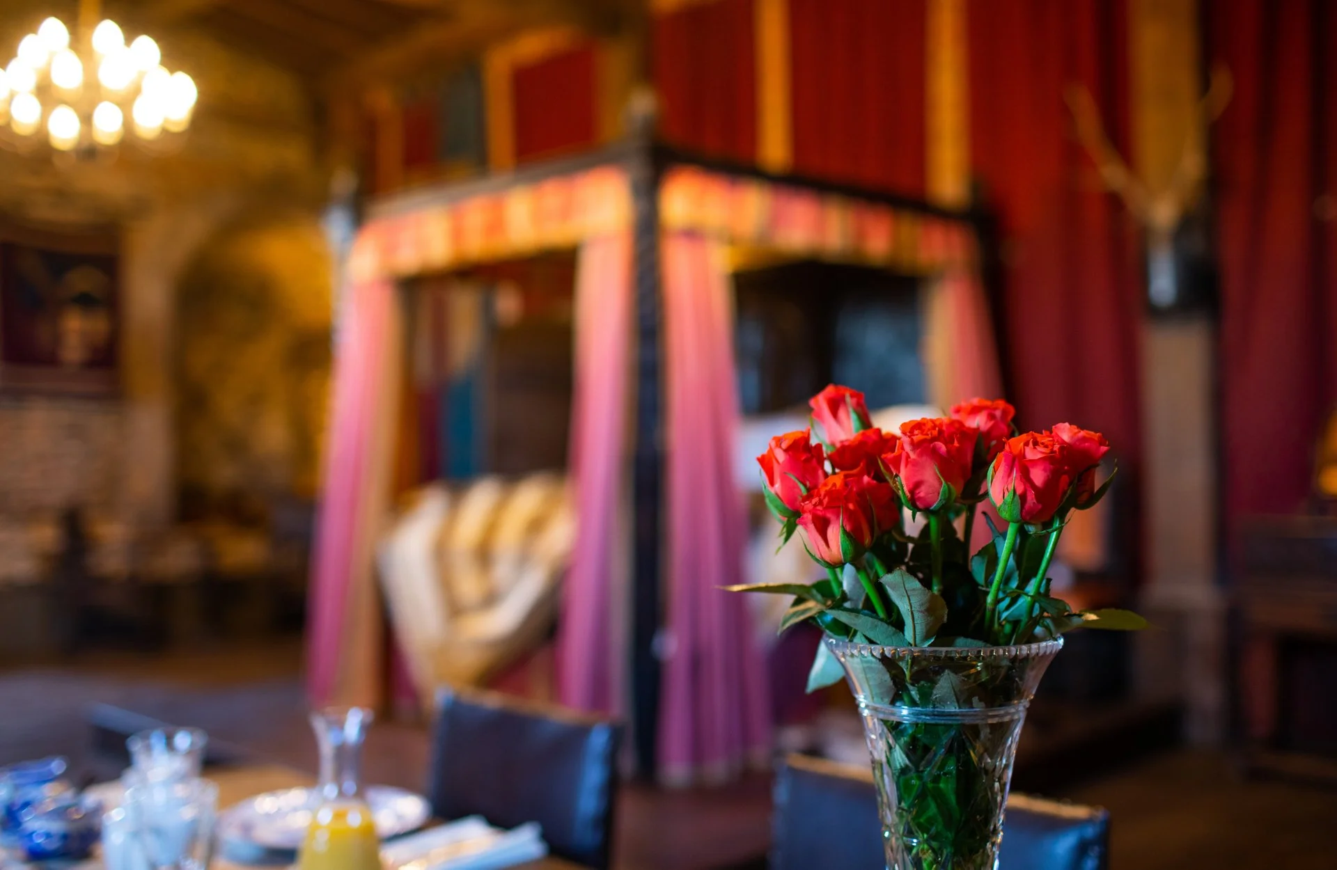 A bouquet of red roses in a glass vase, with a blurred background featuring a medieval-style room, red curtains, a canopy bed, chandelier, and dinner table.