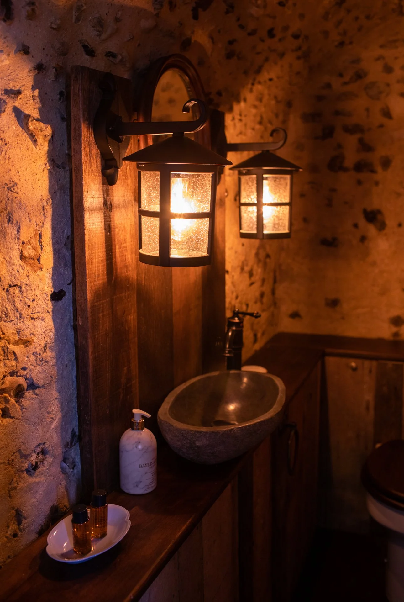 Rustic bathroom with stone walls, two lantern wall lights, a stone basin sink, and a wooden countertop with toiletries.