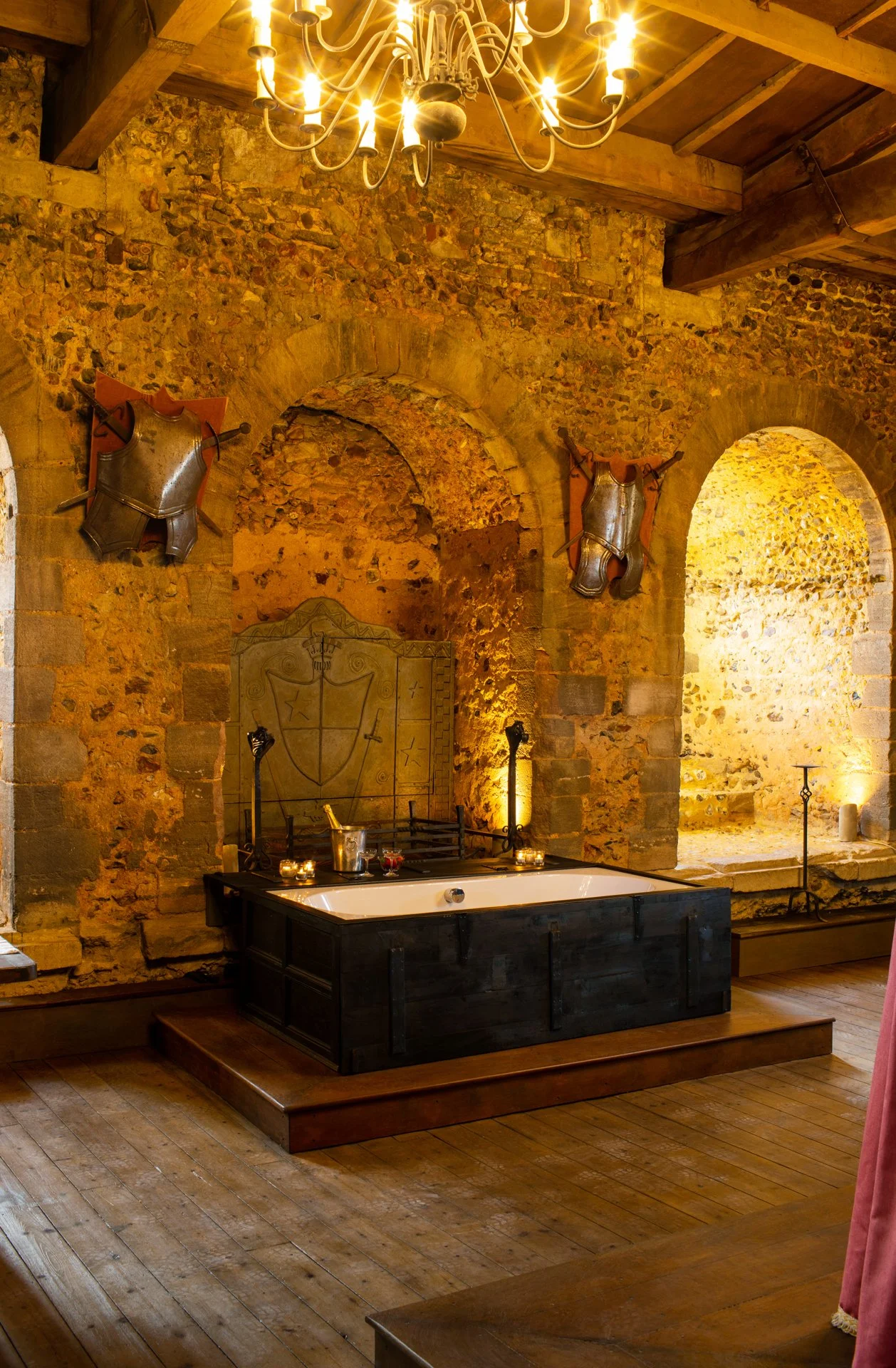 Interior of a medieval-style room with stone walls, a large chandelier, a raised platform with a white bathtub, and decorative armor and shield on the walls.