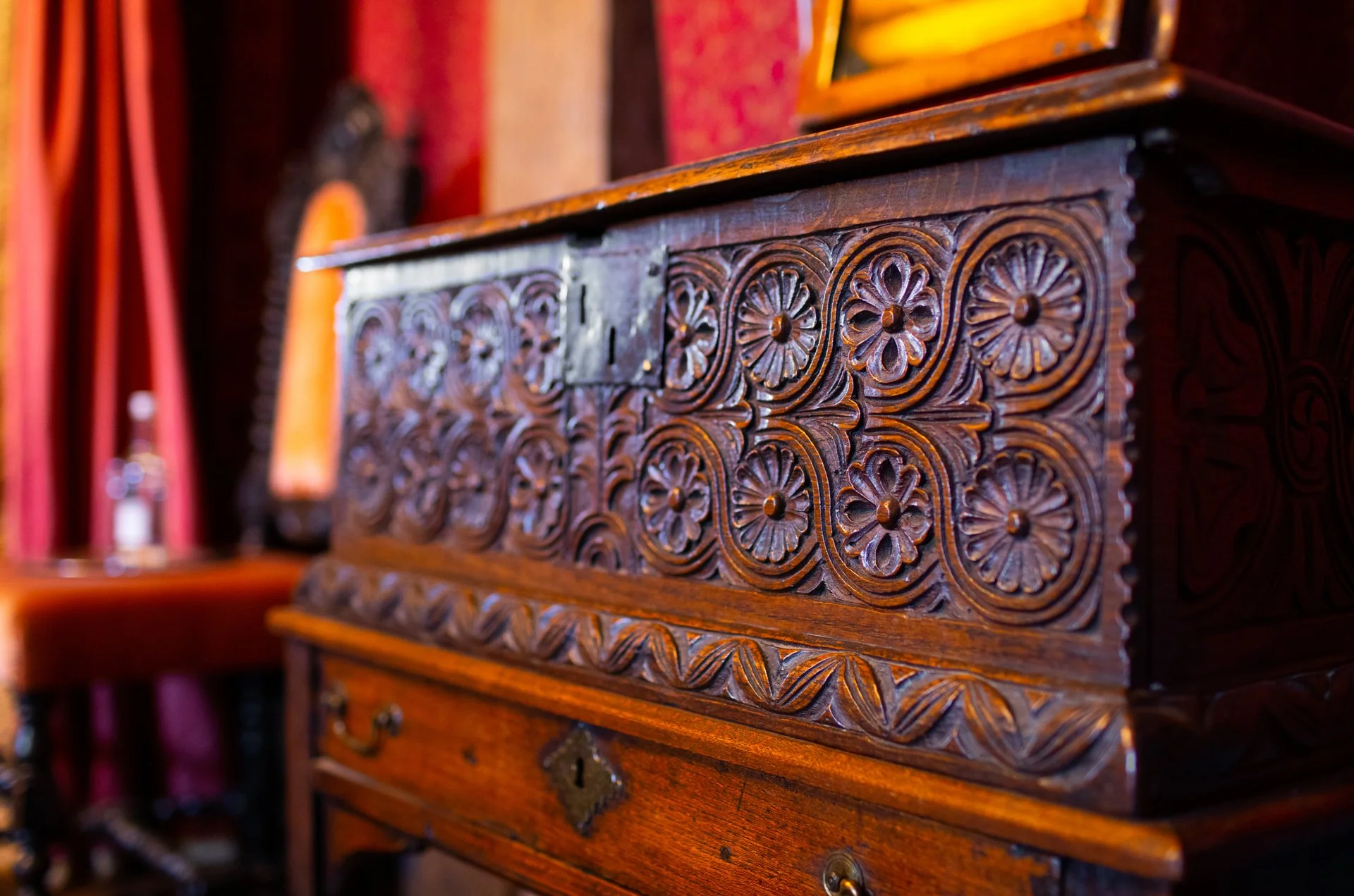 Close-up of an ornately carved wooden chest with floral patterns and metal latch, set against a backdrop of rich red curtains and antique decor.