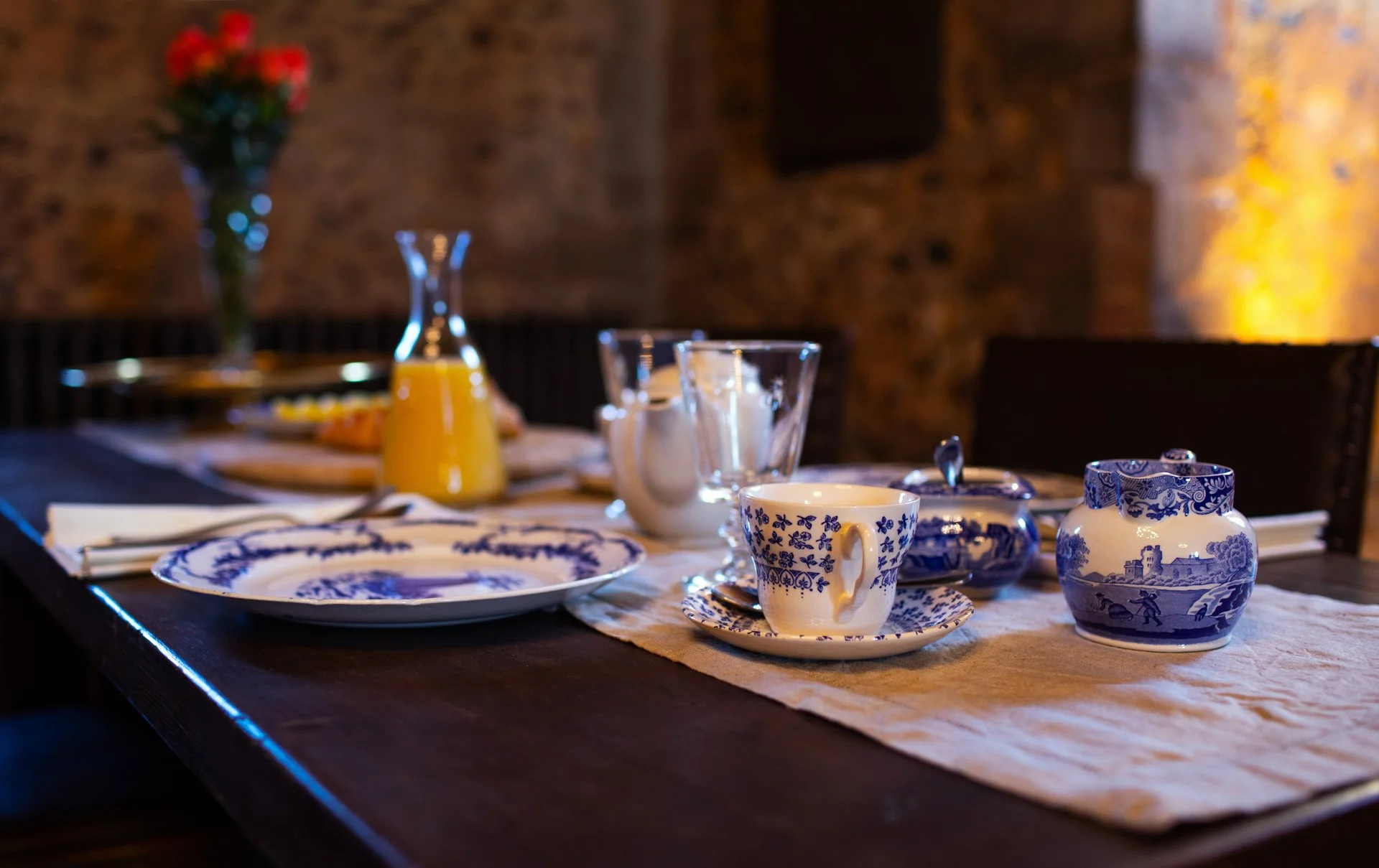 Elegant breakfast table setting with blue and white porcelain teacup, sauce pot, and plate on a tablecloth, a glass pitcher of orange juice, and a bouquet of red flowers in the background.