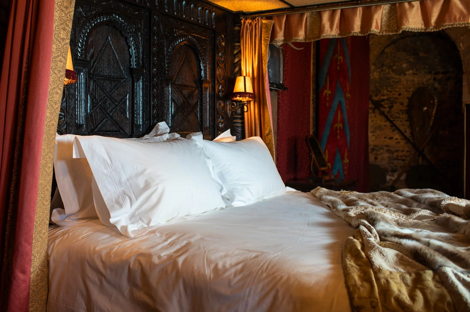 Luxurious bed with white sheets, ornate wooden headboard, velvet and fur coverings, and warm lighting.
