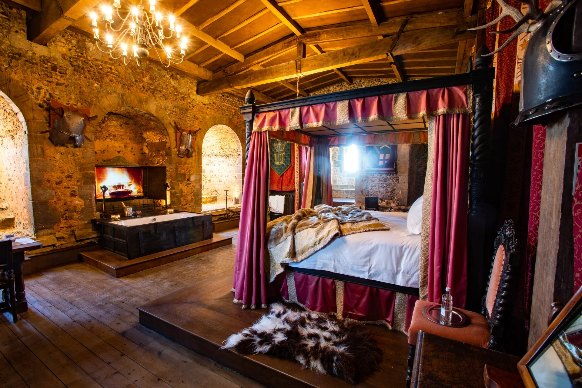 Medieval-style bedroom with stone walls, wooden ceiling, chandelier, four-poster bed with red drapes, animal hide rug, fireplace with firewood, bathtub, and medieval decor.