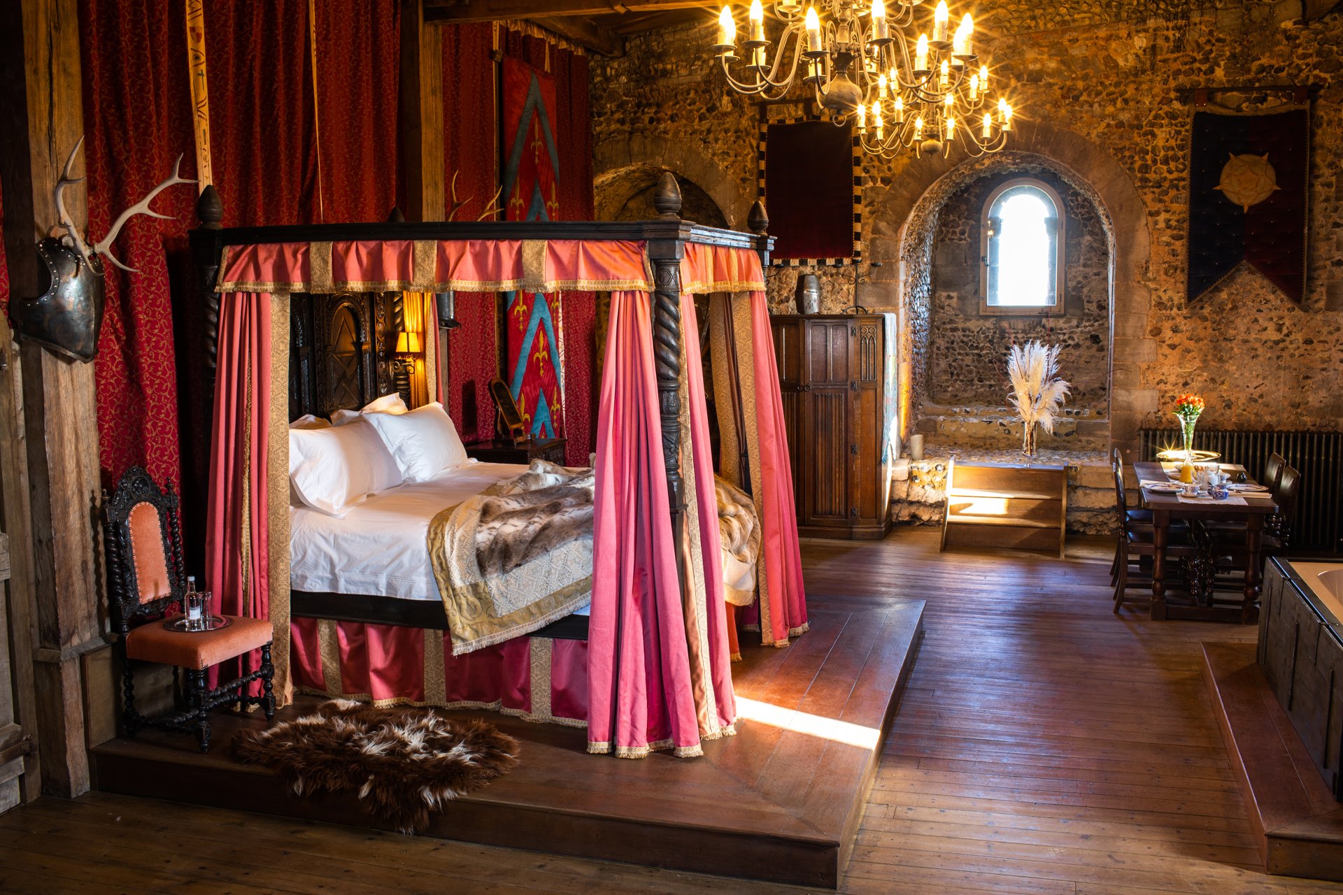 Luxurious medieval-themed bedroom with a canopy bed, stone walls, antique furniture, and a chandelier.