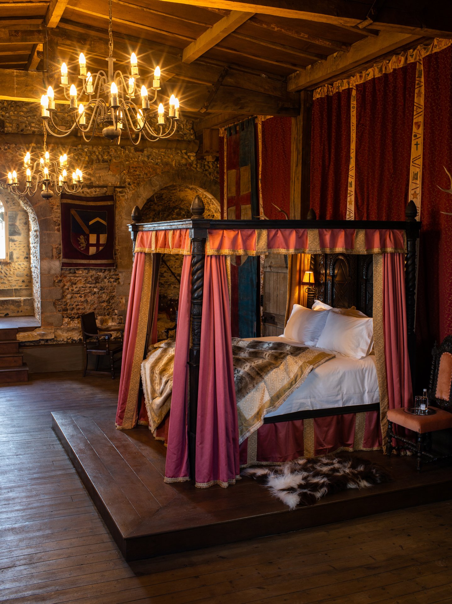 Medieval-style bedroom with a canopy bed, draped in rich fabrics, surrounded by stone walls, elegant chandeliers, and wooden floors.