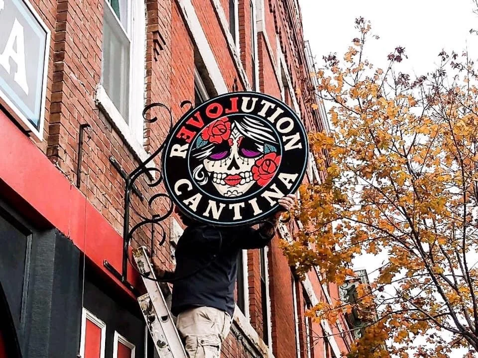 Person installing a circular sign for 'Revolution Cantina' on a brick building wall, with a tree and autumn leaves in the background.