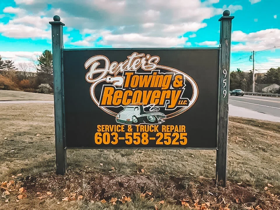 Sign for Dexter's Towing & Recovery, offering service and truck repair, with phone number 603-558-2525, located by a road with cars, trees, and partly cloudy sky in the background.