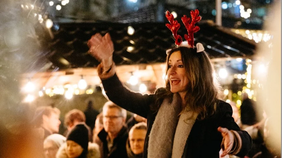 Woman wearing reindeer antler headband and smiling, Christmas lights in background, festive gathering.