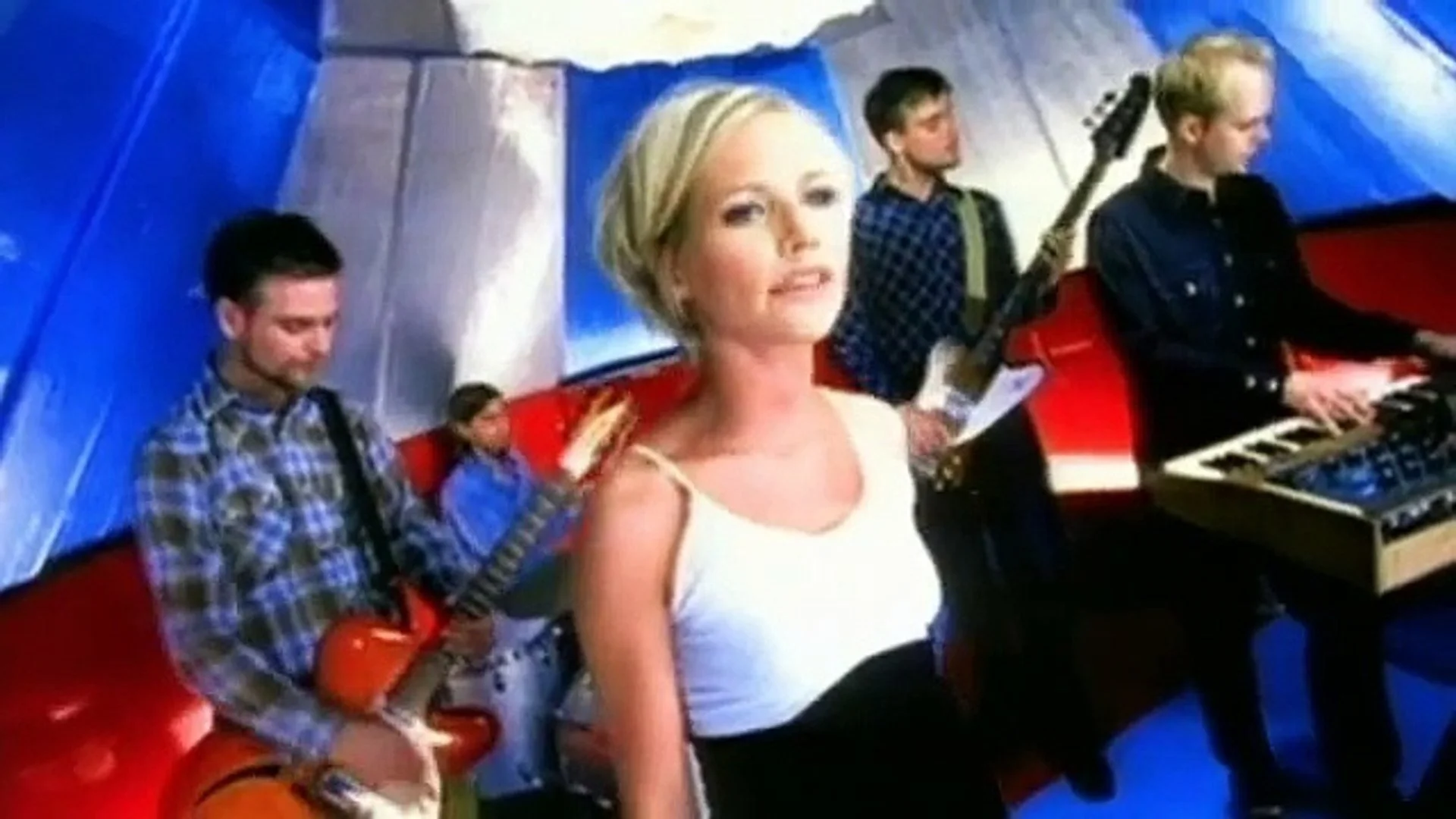 A woman with blond hair and a white top sitting on a red bench in front of a blue and white background, with musicians playing instruments behind her, including a man playing the keyboard and another man playing the guitar.