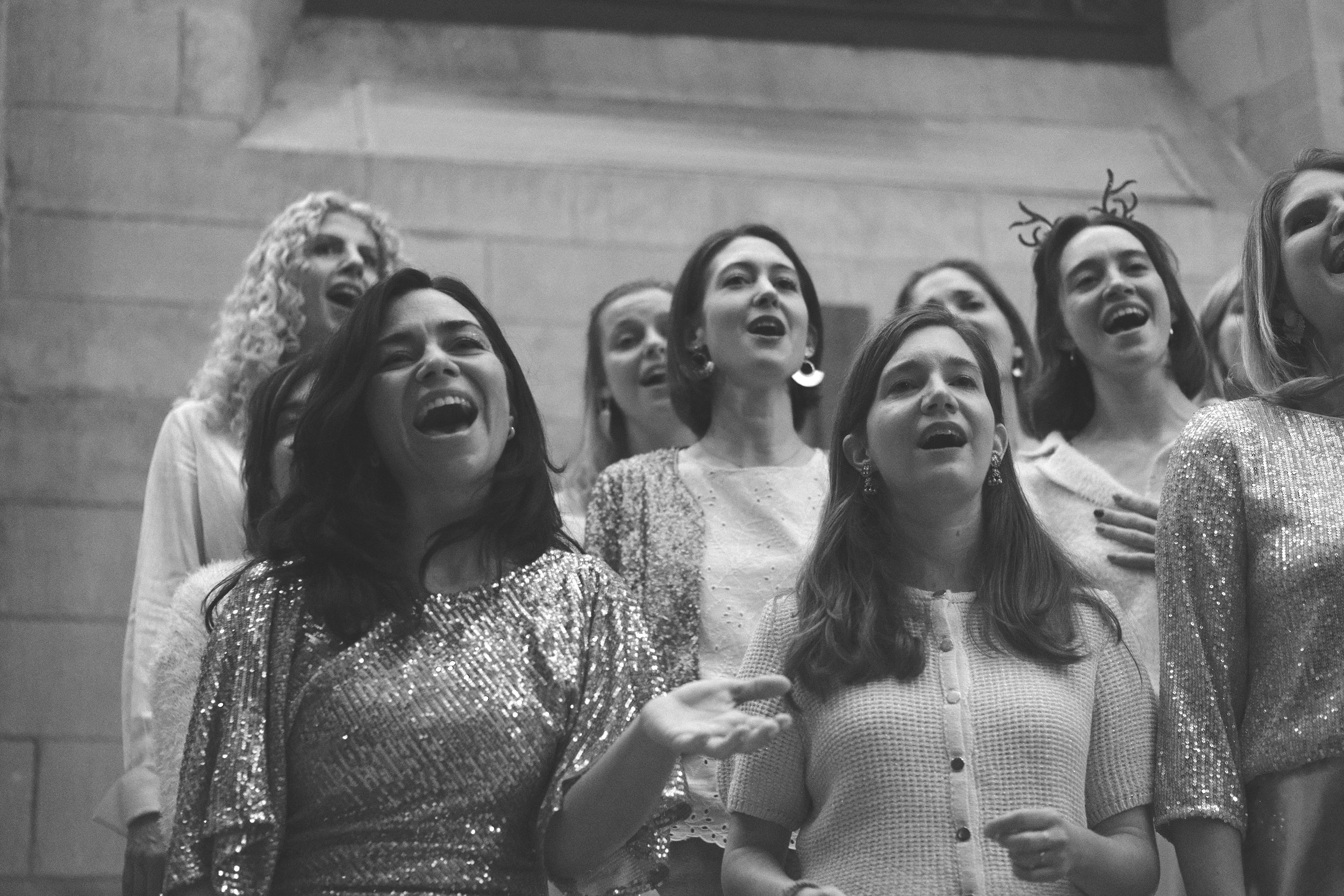Group of women singing Christmas carols, some wearing holiday accessories like reindeer antlers, in a festive indoor setting.