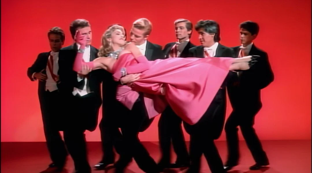 A woman in a pink dress is being carried by seven men in tuxedos against a red background.