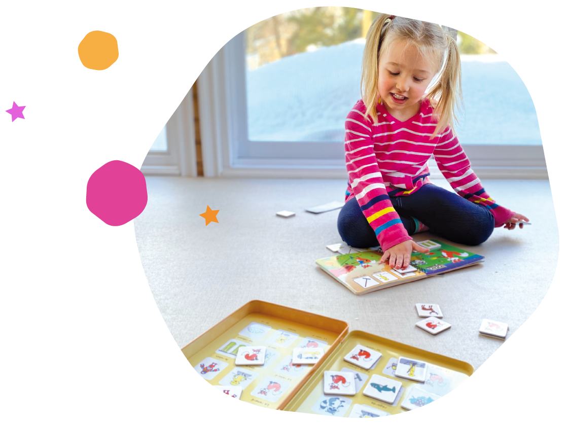Une petite fille joue avec un jeu de société ou des cartes illustrées à l'intérieur, près d'une grande fenêtre laissant entrer la lumière naturelle.