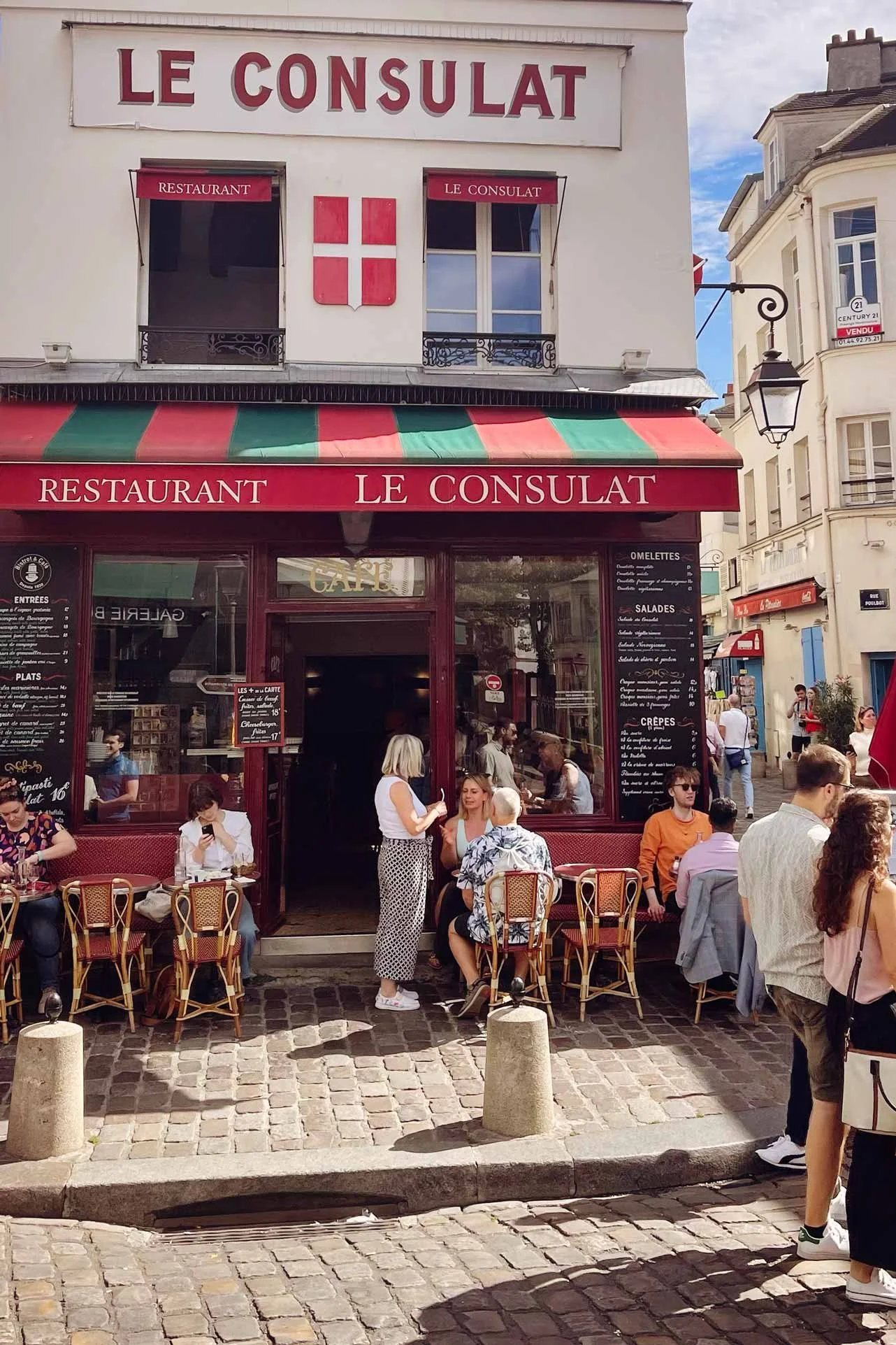 terras restaurant le consulat Montmartre Parijs met mensen op het terras die eten, kletsen en rondkijken en op hun telefoon kijken. groen rode luifel met witte gevel en rode letters