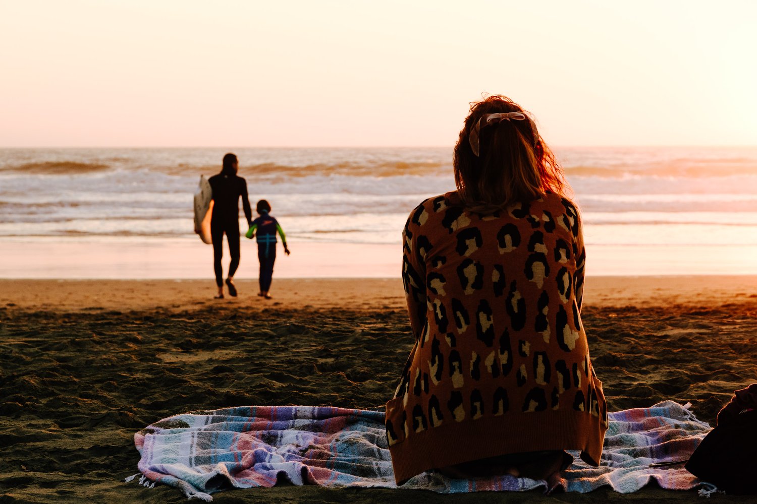 Mom sits on a beach towel watching her son and a surf instructor walk towards the water at Ocean Beach.