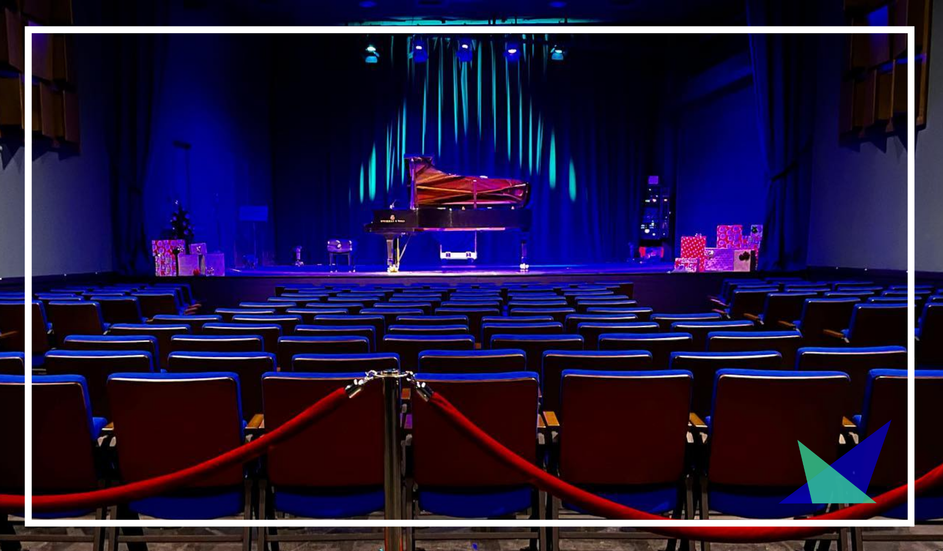 Empty theater with a grand piano on stage, decorated with Christmas presents and a Christmas tree, with blue lighting.