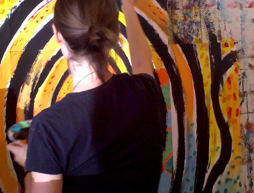 Woman standing at a wall painting large yellow and black spirals on paper