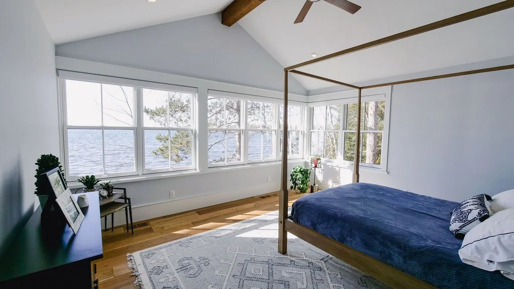 Bedroom with large windows overlooking a lake, a wooden four-poster bed with blue bedding, and a dark dresser with picture frames and plants.