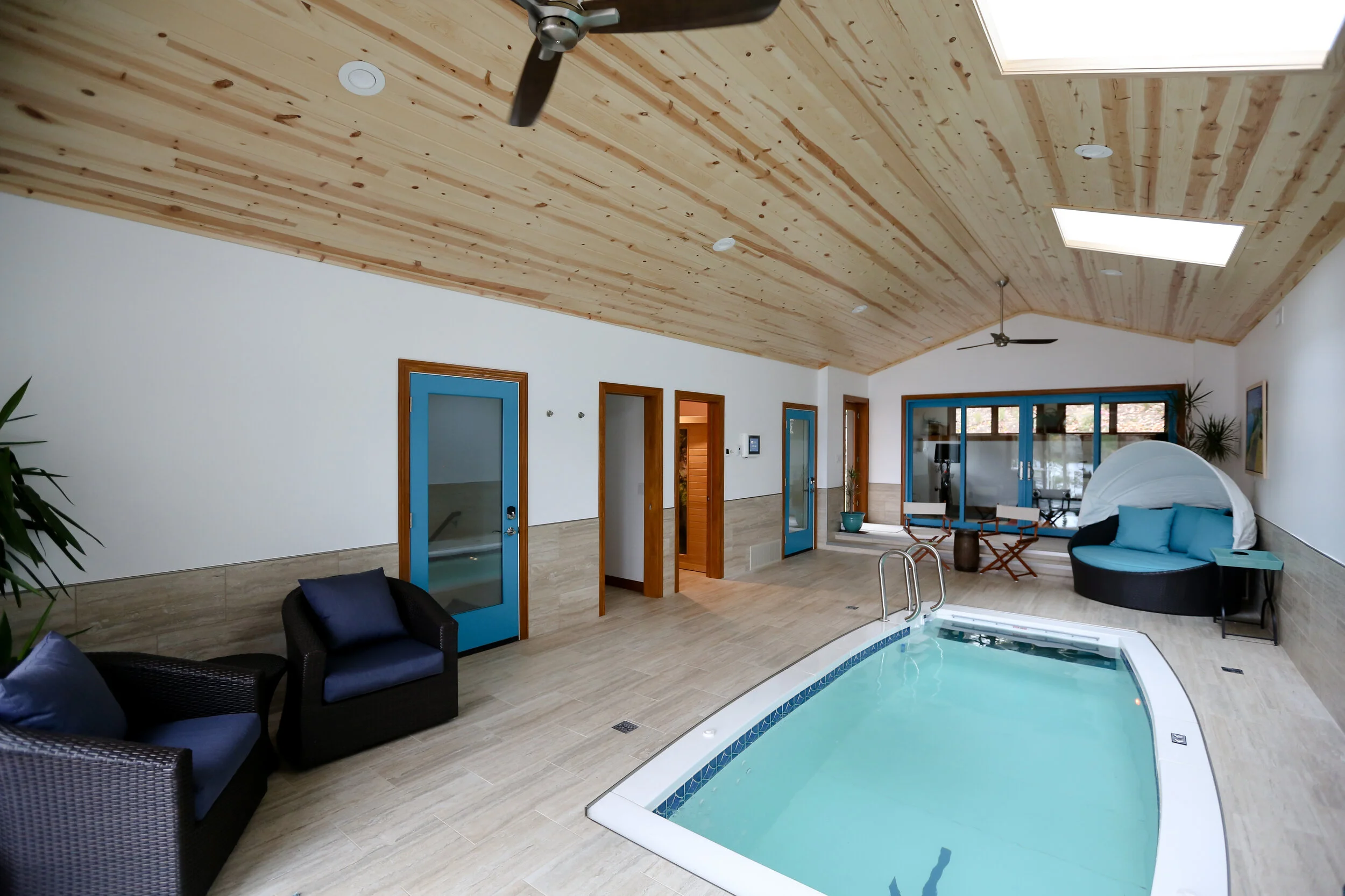 Indoor pool area with wooden ceiling, ceiling fans, skylights, and beige tiled floor. There are blue-framed glass doors, black wicker lounge chairs with blue cushions, a round daybed with blue pillows and a white canopy, and potted plants.