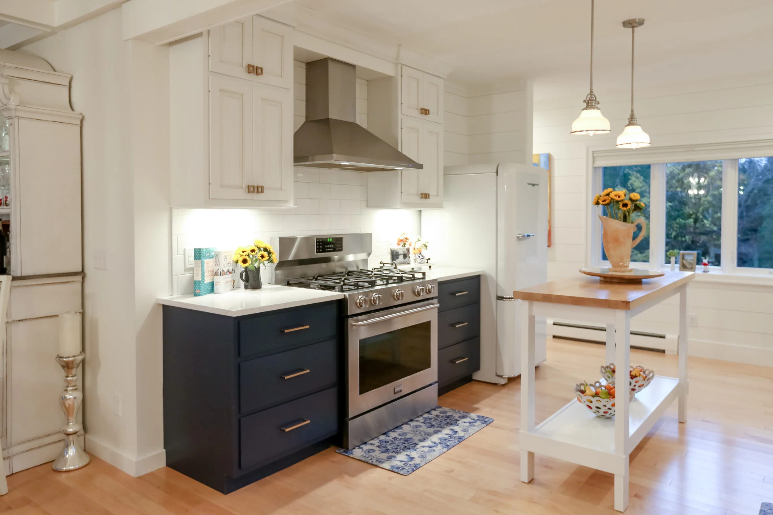 Kitchen with white cabinets, stainless steel oven and range, white countertop, blue lower cabinets, white subway tile backsplash, white refrigerator, wooden kitchen island with a vase of sunflowers, hardwood floors, window with a view of greenery, and pendant lights.