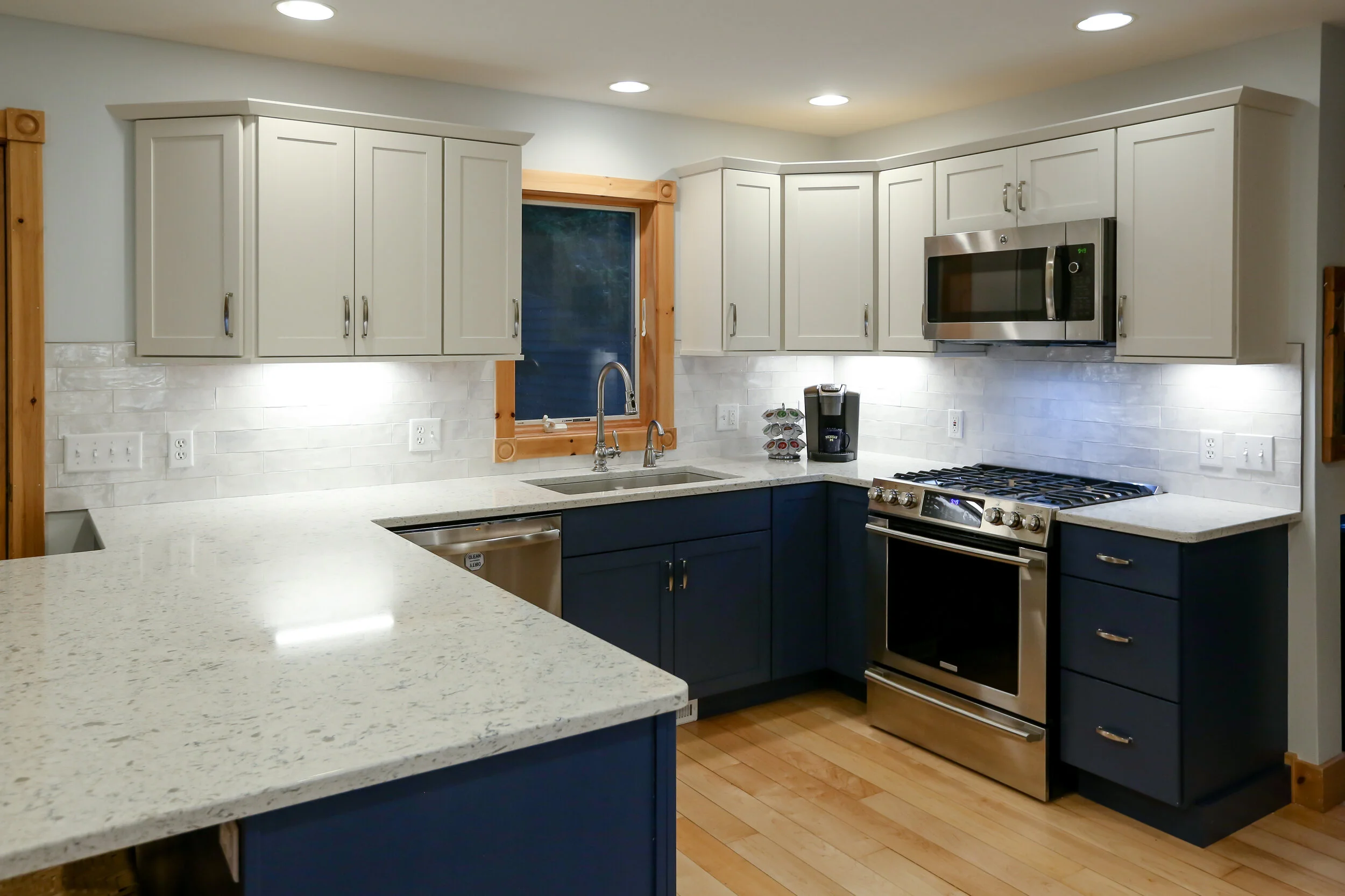 Modern kitchen with white upper cabinets, blue lower cabinets, a white speckled countertop, stainless steel appliances including a stove and microwave, a window above the sink, and a wooden floor.