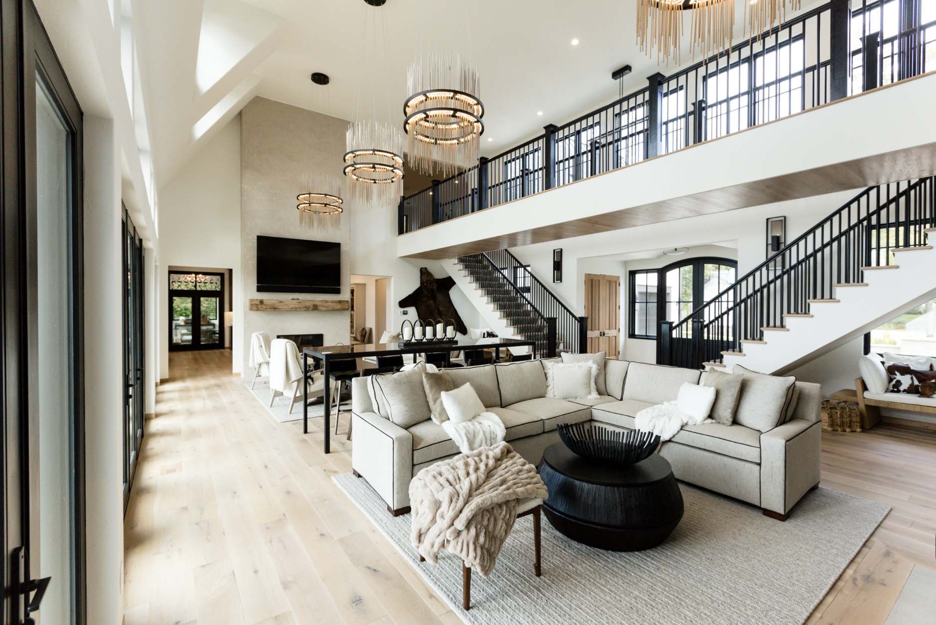 Spacious modern living room with beige sectional sofa, light wood flooring, black metal staircase, large windows, and contemporary lighting fixtures