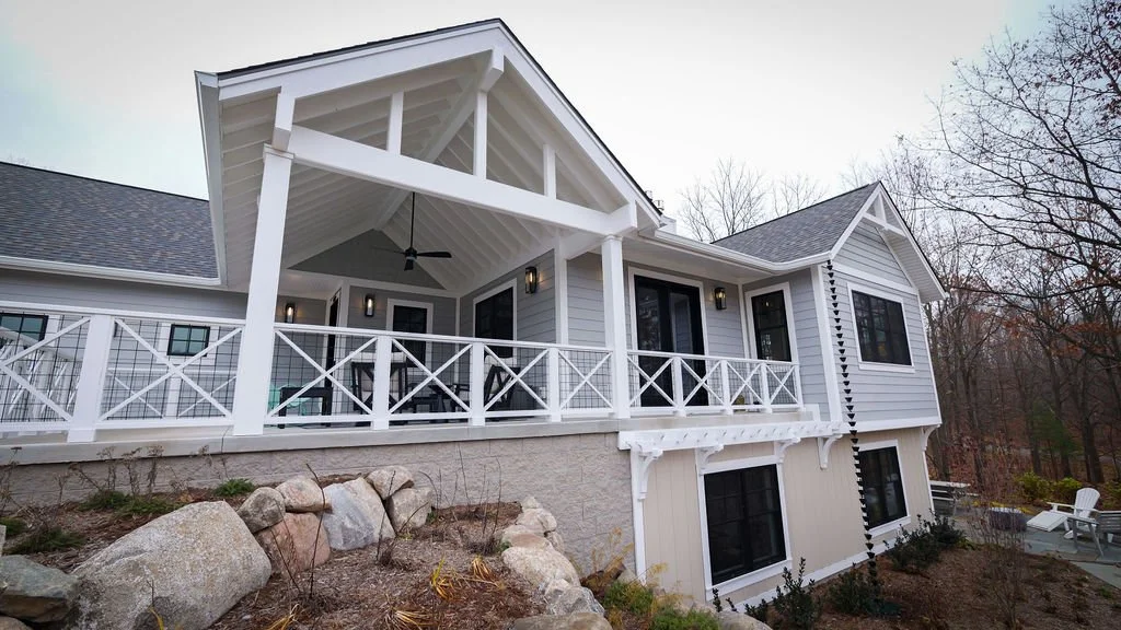 Front view of a modern house with a large covered porch, white siding, and black window frames, situated on a hillside with rocks and trees in the background.