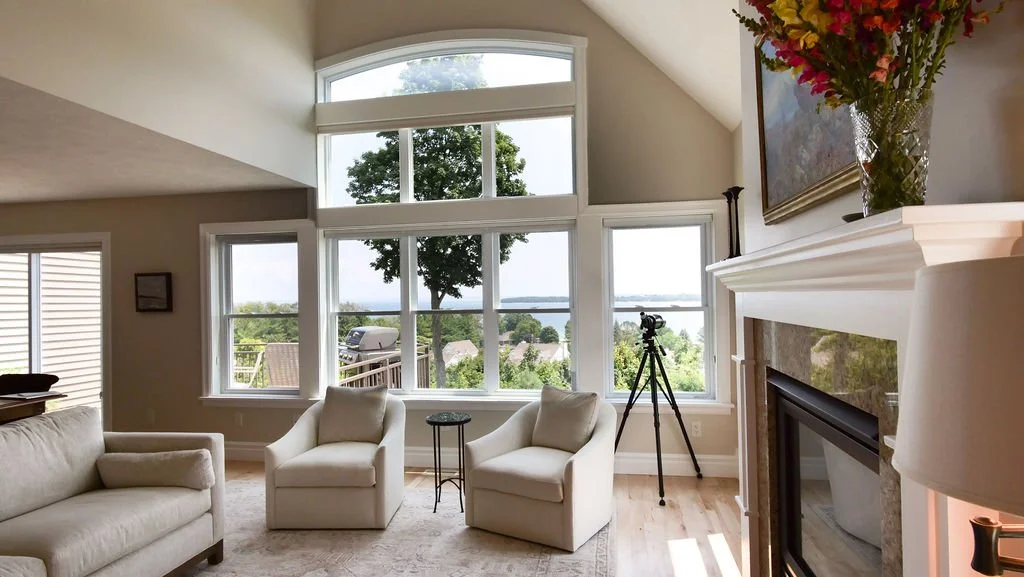 Living room with large windows showing trees and water, beige furniture, a fireplace with a mantle holding a vase of flowers, and a telescope pointing outside.